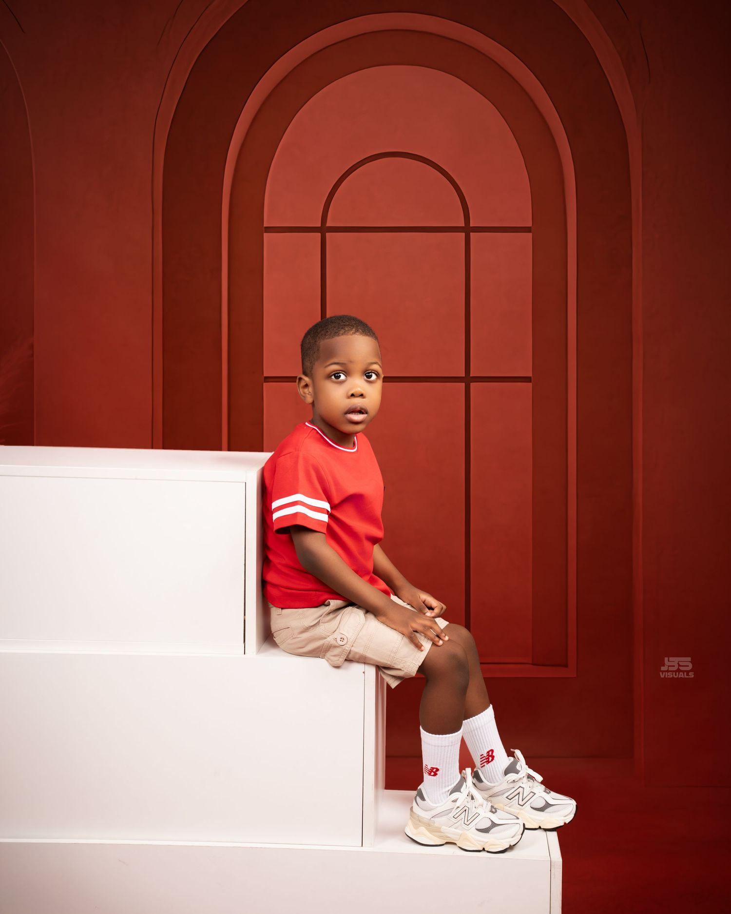 Young boy in red shirt and New Balance sneakers sits on white steps against a bold red arched backdrop.