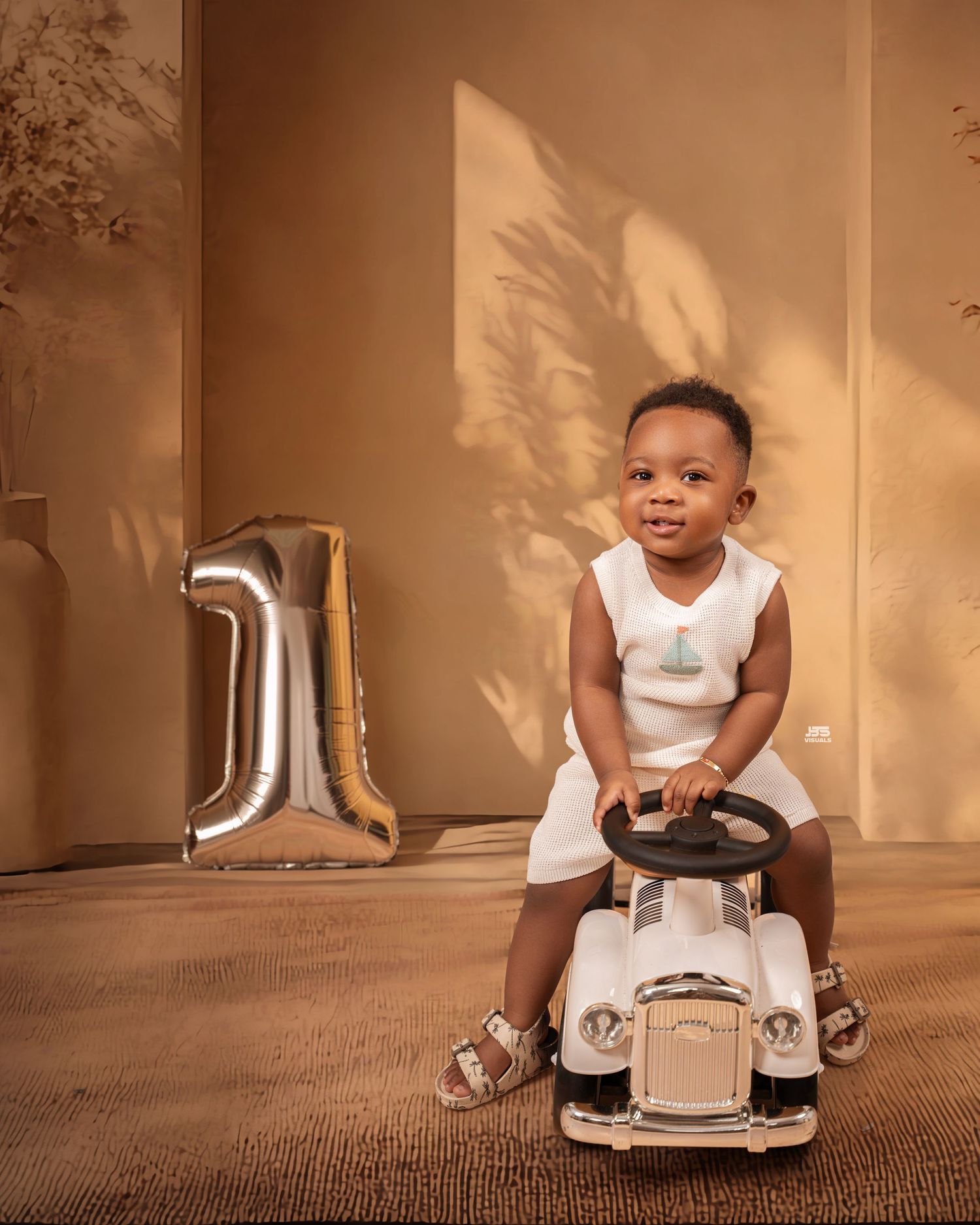 Smiling baby sitting on toy car beside silver number 1 balloon, celebrating first birthday in warm beige studio setting.
