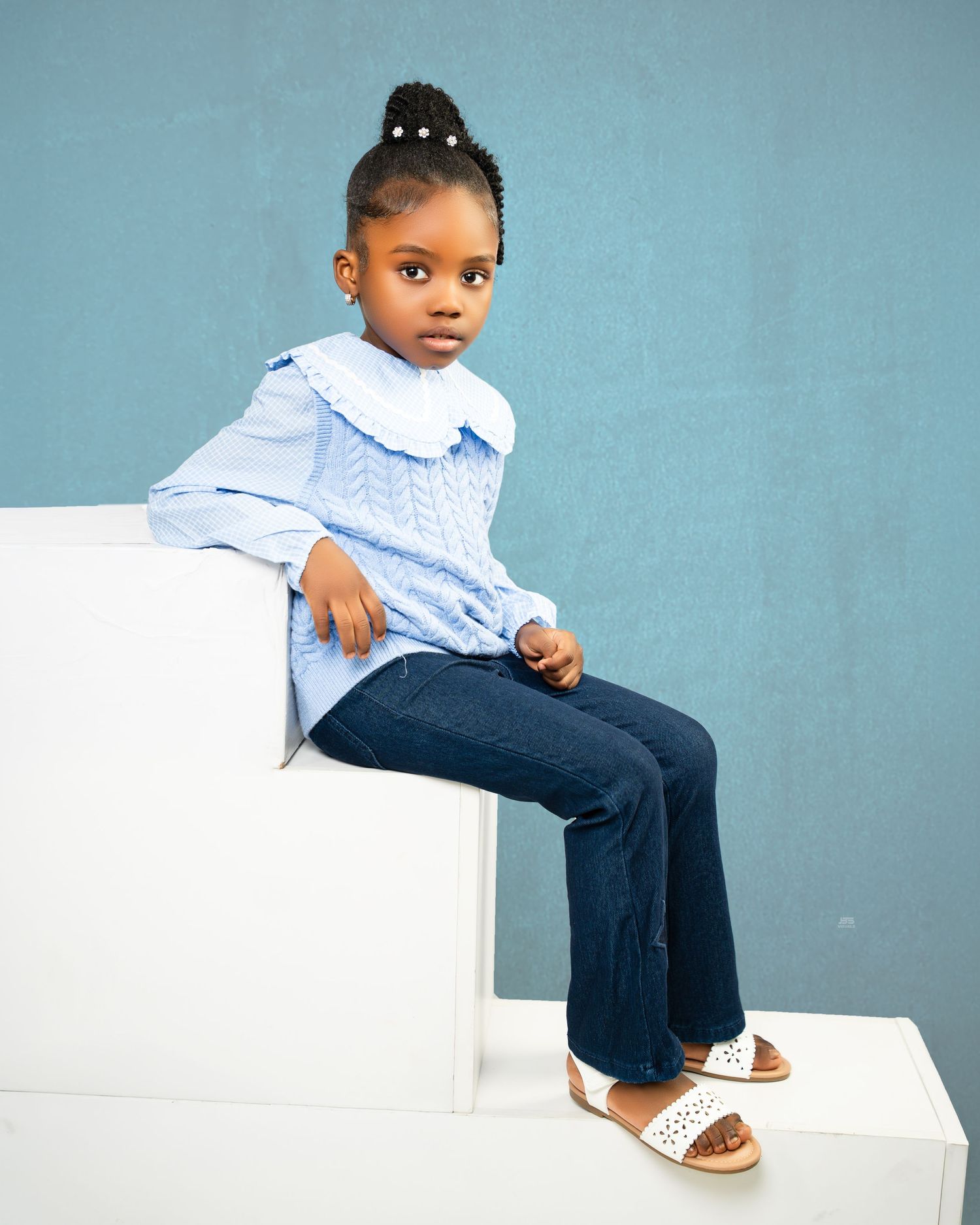 Young girl in blue ruffled blouse and jeans sitting on white cube against blue background.