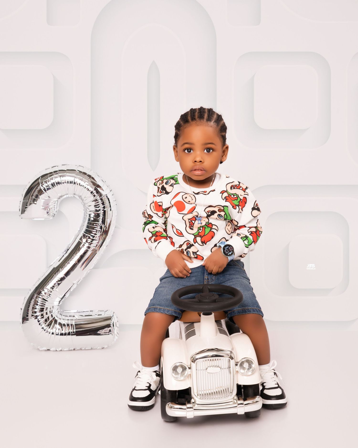 Toddler boy in graphic sweatshirt sits on white ride-on car beside silver number 2 balloon for birthday shoot.