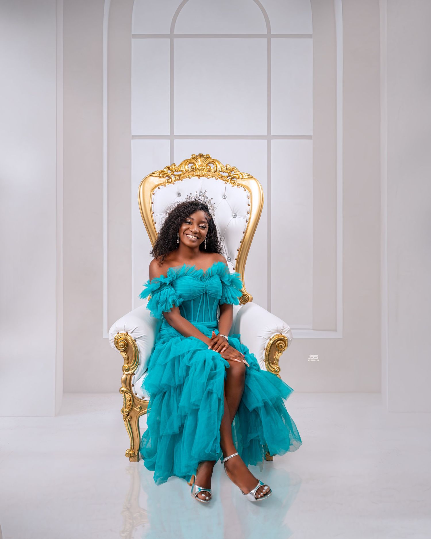 Woman in teal ruffled off-shoulder gown seated on ornate white and gold throne chair in bright white studio.