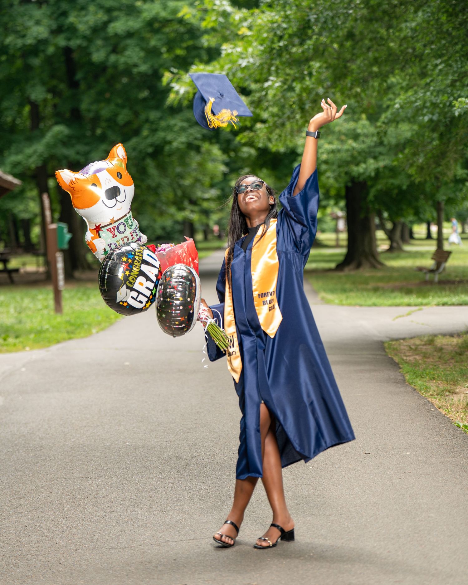 Joyful graduate tossing cap in air while holding balloons in park.