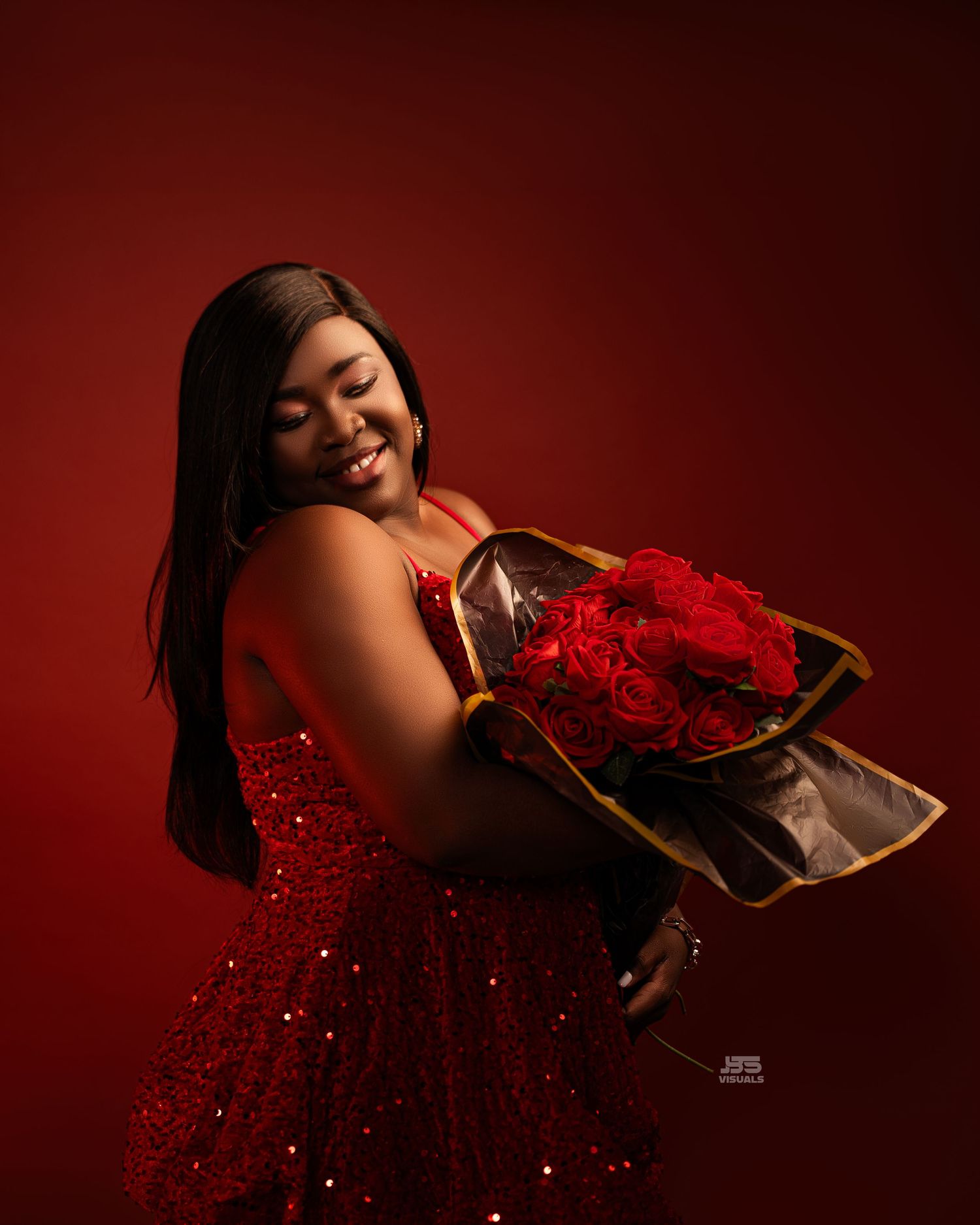 A smiling woman in a red sequin dress holds a bouquet of red roses against a deep red studio background.