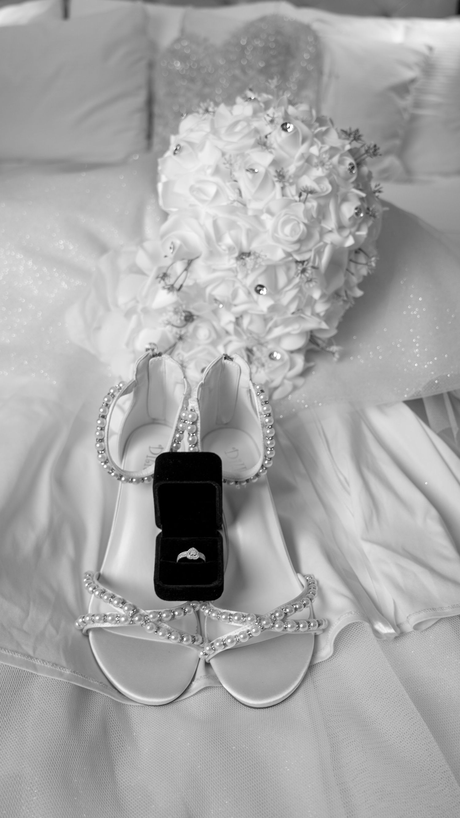 Black and white flat lay of bridal accessories including sandals, diamond ring box, and white rose bouquet.
