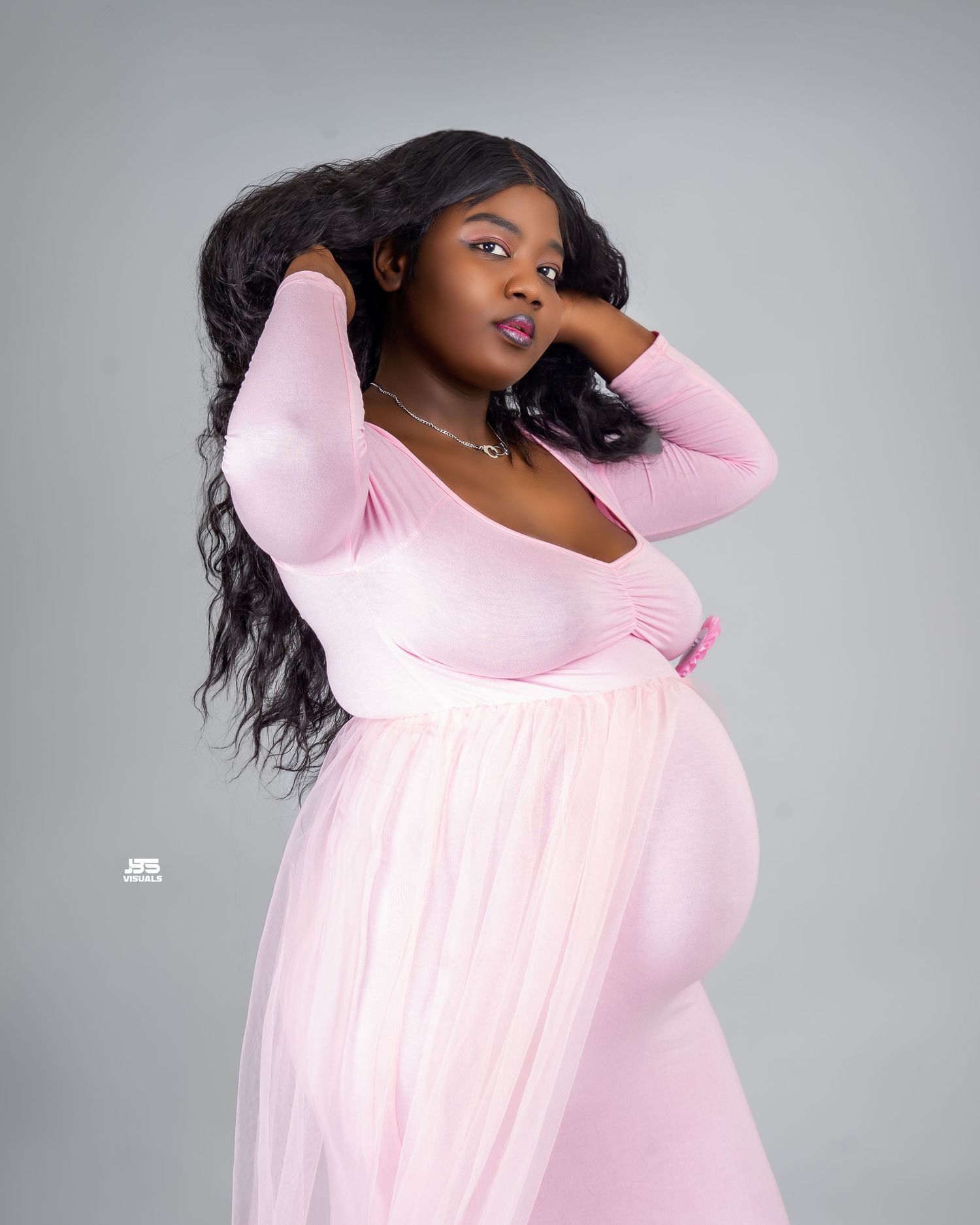 Pregnant woman in pink tulle maternity gown poses elegantly in professional studio photo shoot.