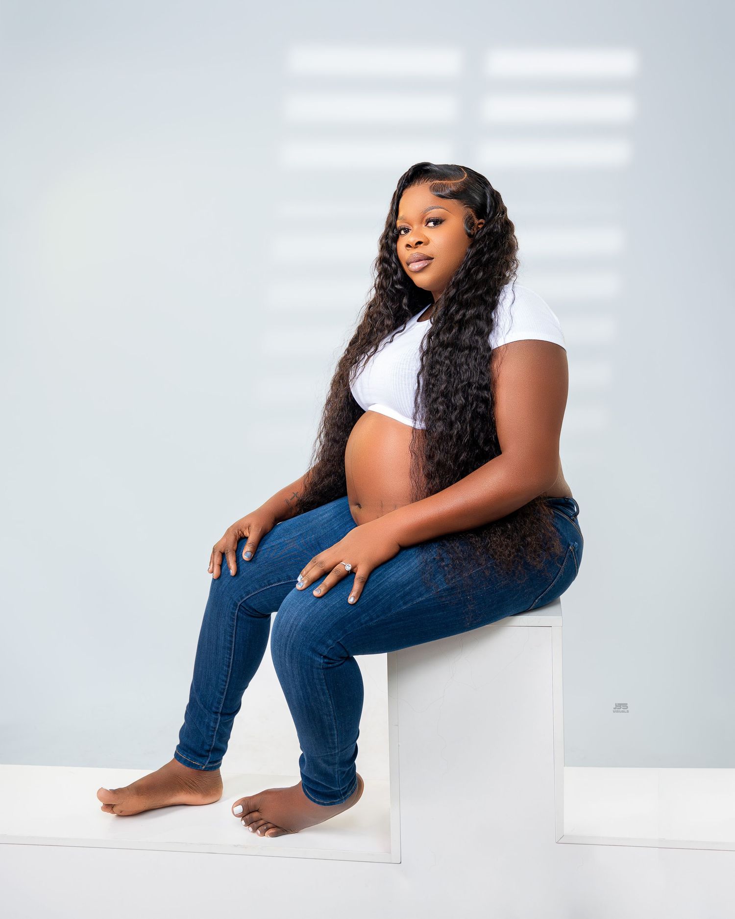 Pregnant woman with long curly hair sits on white cube in casual maternity photo wearing jeans.