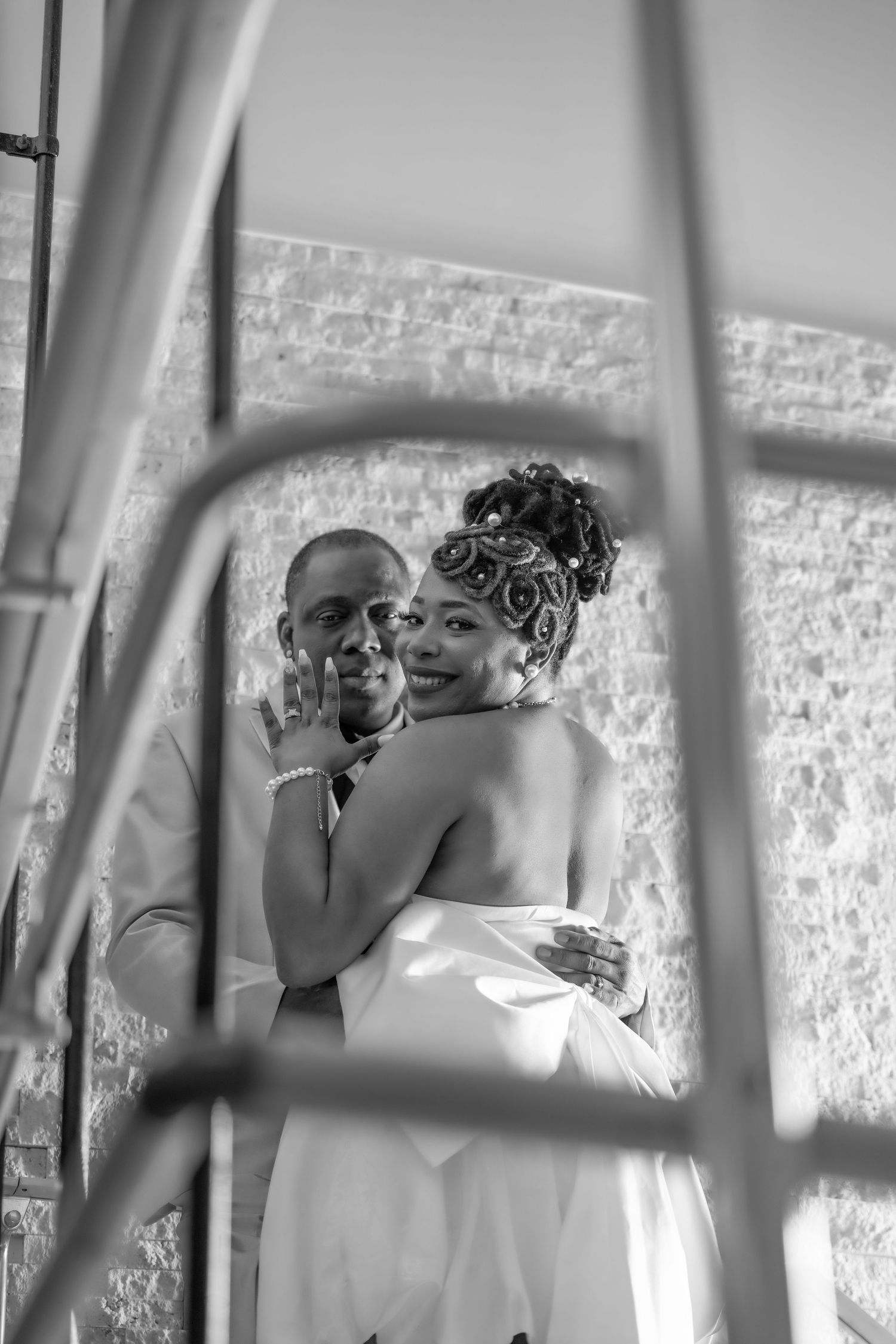 Black and white photo of a smiling bride and groom embracing, viewed through decorative metal railings.