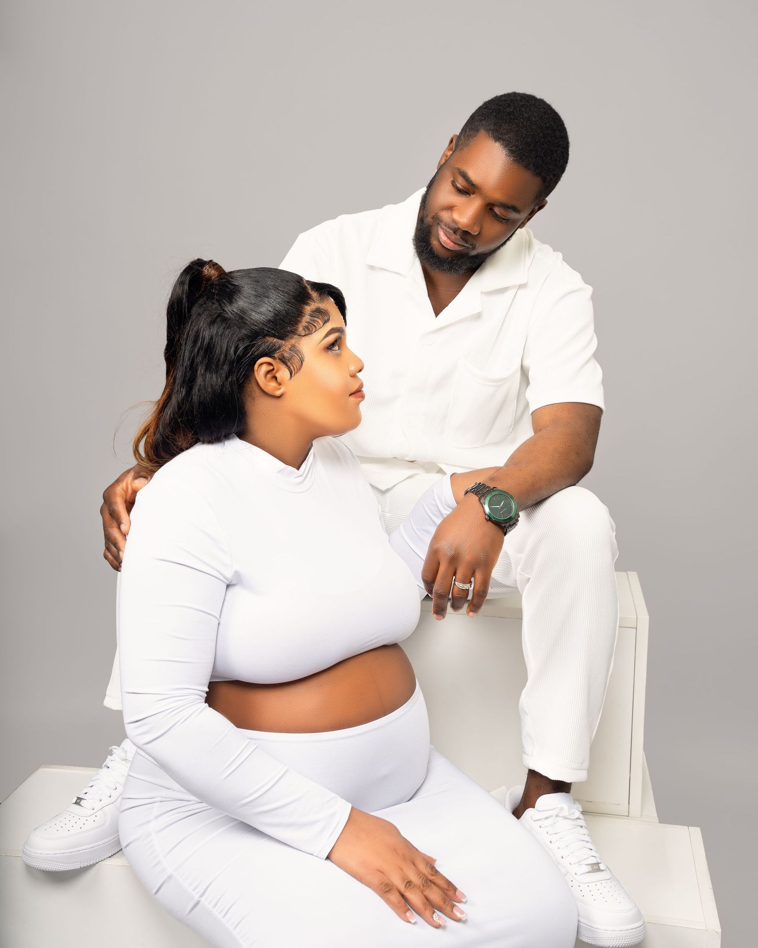 Expecting couple in matching white outfits sharing a tender moment in a studio portrait.