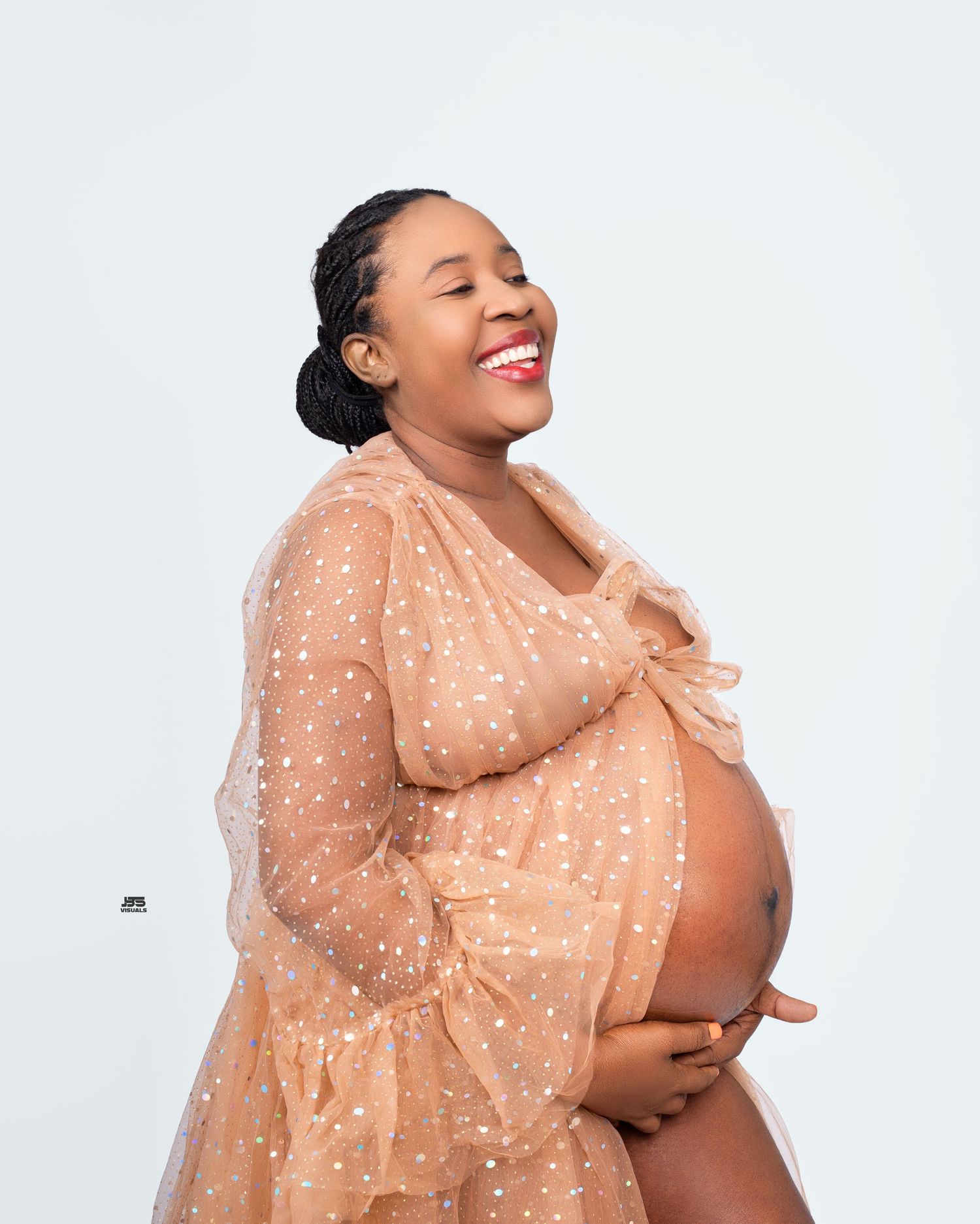 Joyful pregnant woman in gold polka dot sheer robe cradling her baby bump on white backdrop.