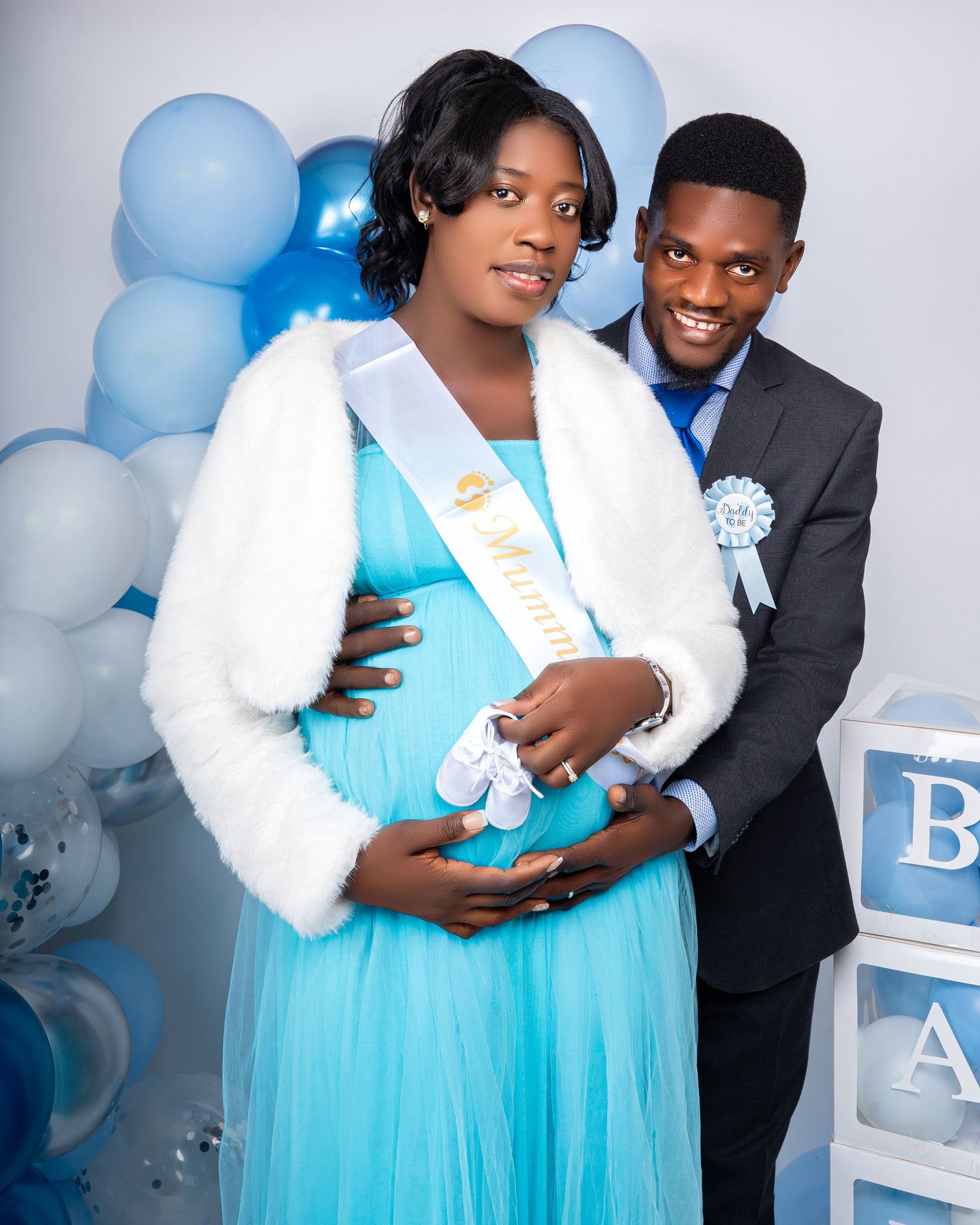 Couple posing at baby boy maternity shoot with blue balloons, BABY boxes, and white fur jacket.