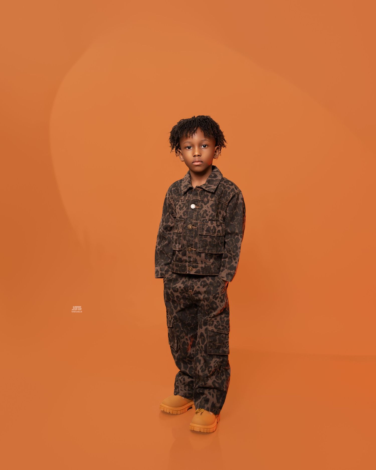 Young boy in brown leopard print jacket and pants stands against an orange background in a studio portrait.
