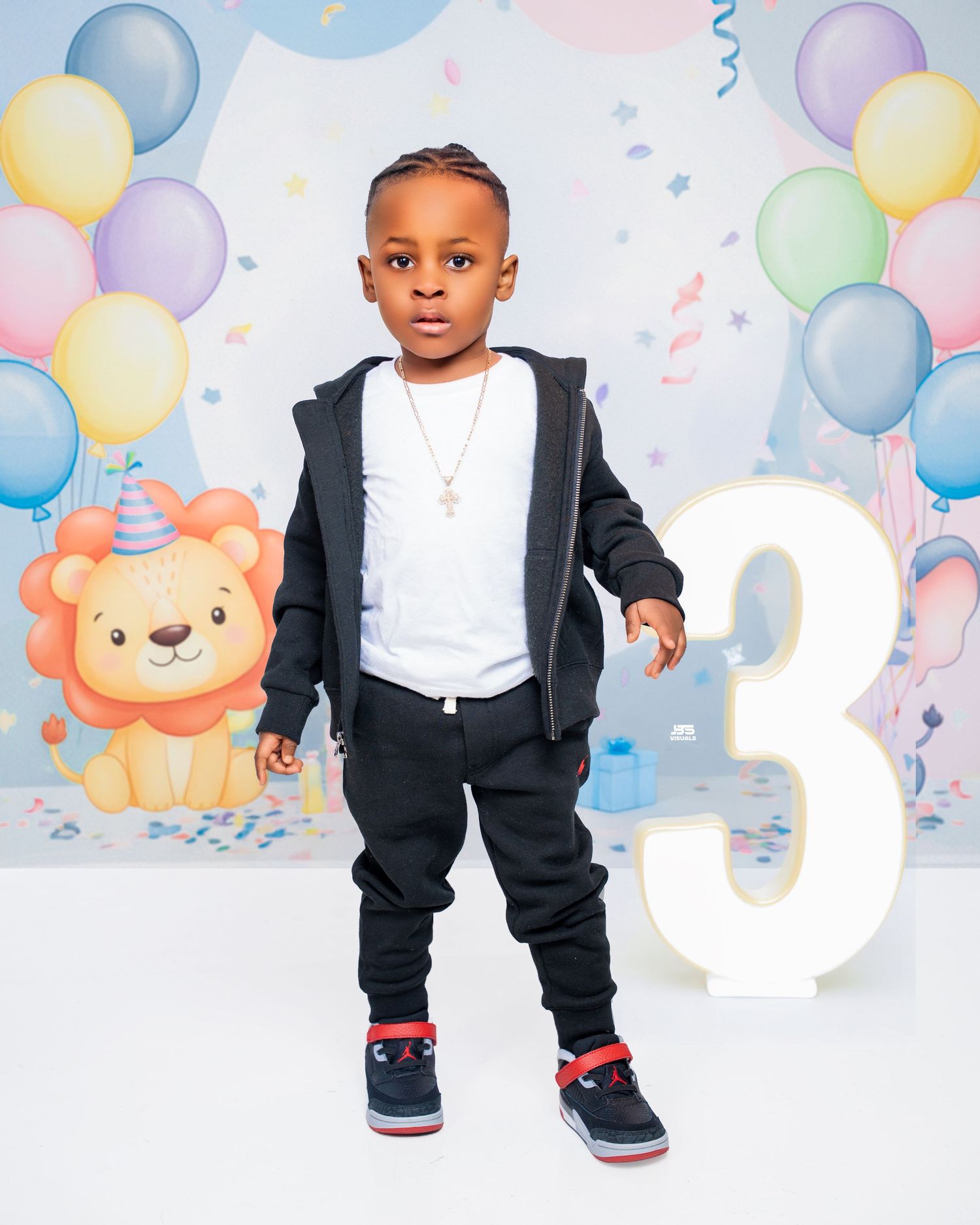 Young Black boy in black tracksuit posing with number 3 sign at colorful balloon birthday backdrop.