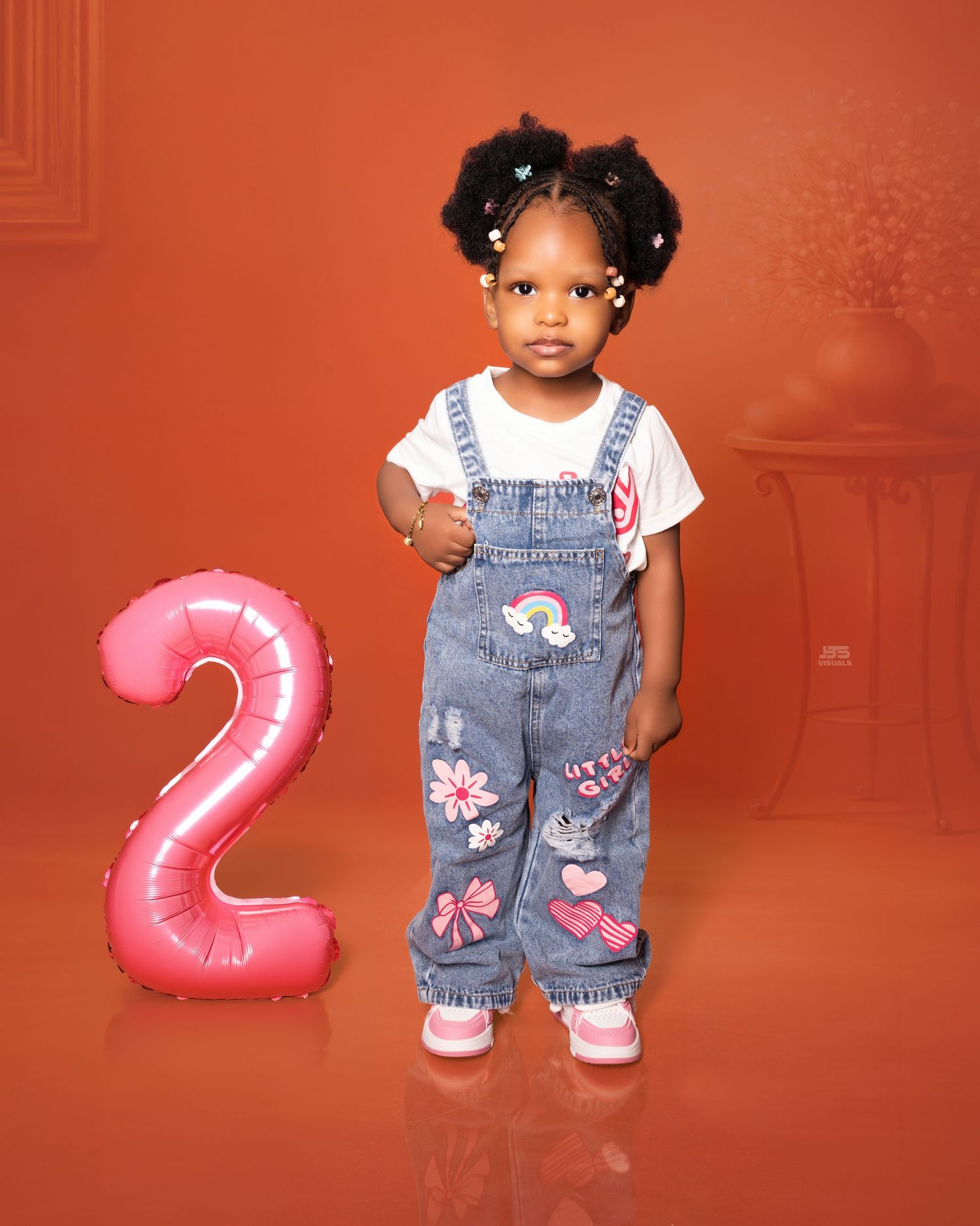 Cute toddler girl in denim overalls stands beside a pink number 2 balloon for her 2nd birthday photos.