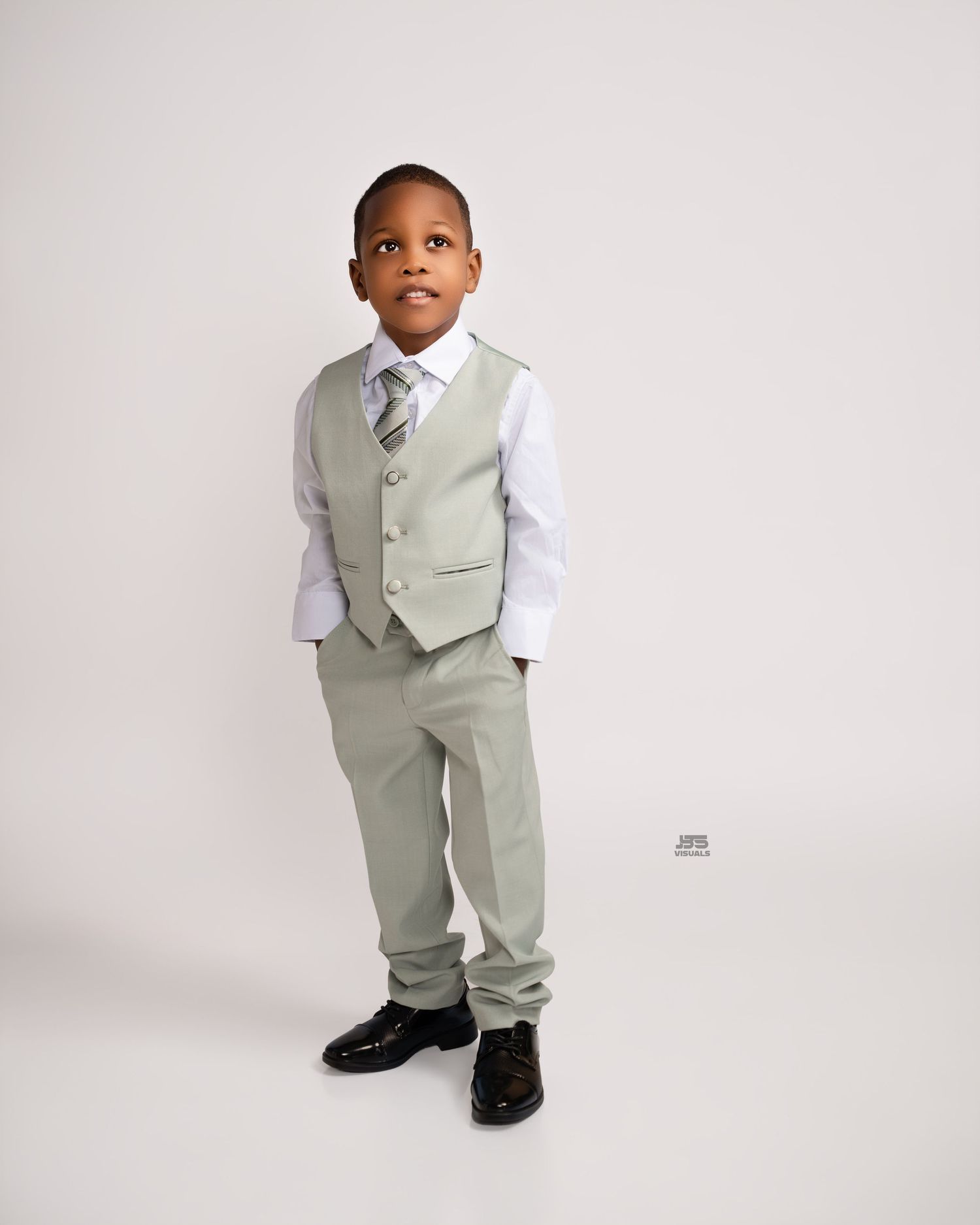 Young boy in sage green vest suit with white shirt, tie, and dress shoes poses against white background.