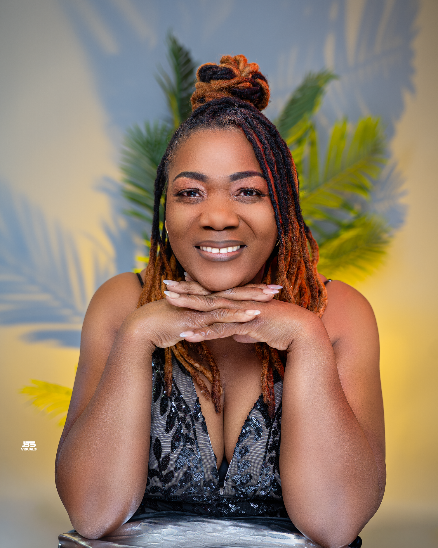 Smiling woman with orange dreadlocks in a black dress posing with hands clasped at a tropical studio.