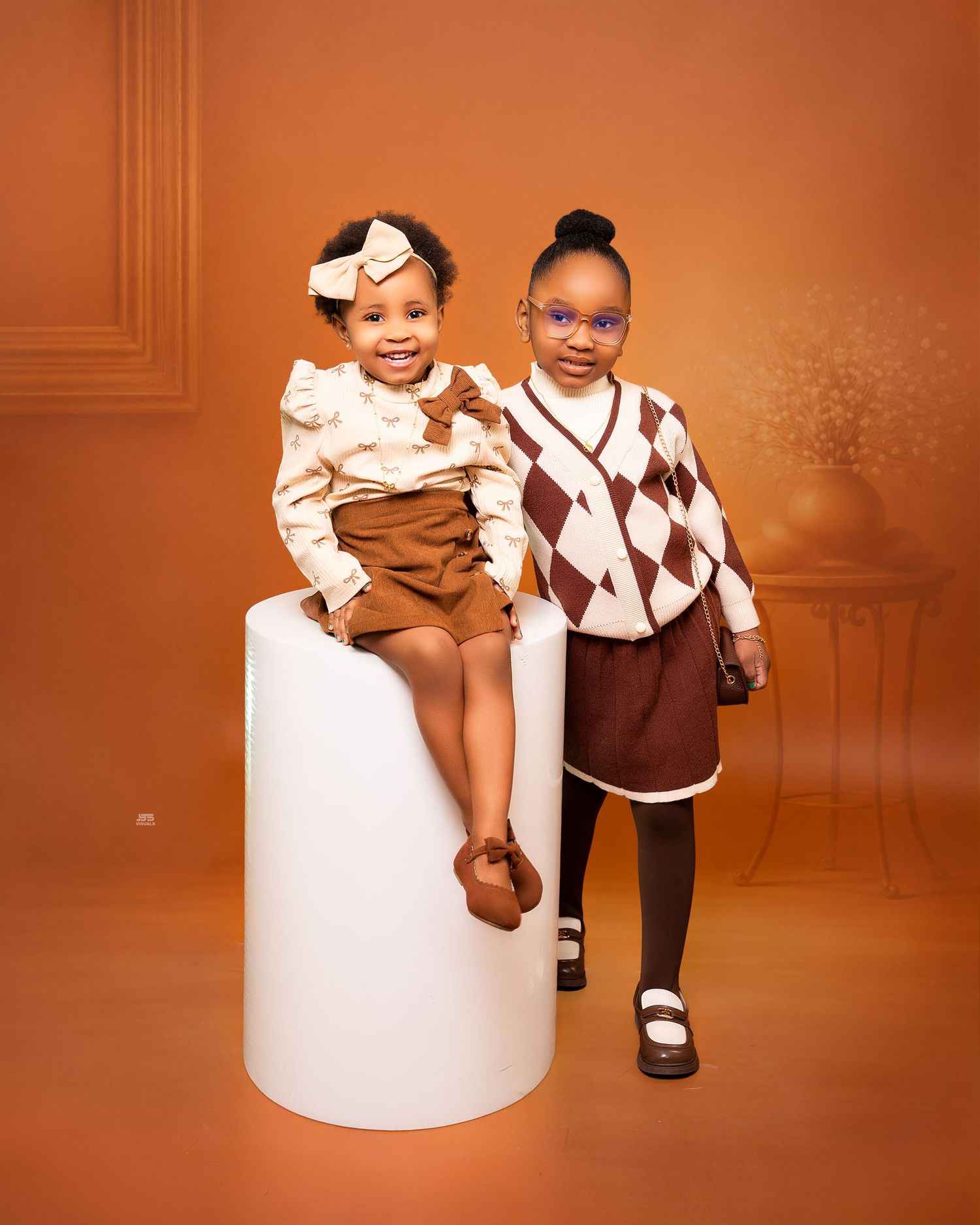 Two young girls posing together in coordinated brown outfits against warm orange background.