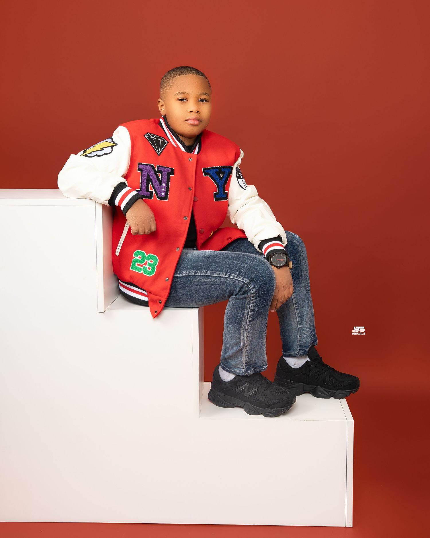 Young boy in NY varsity jacket and jeans sitting on white steps against red background.