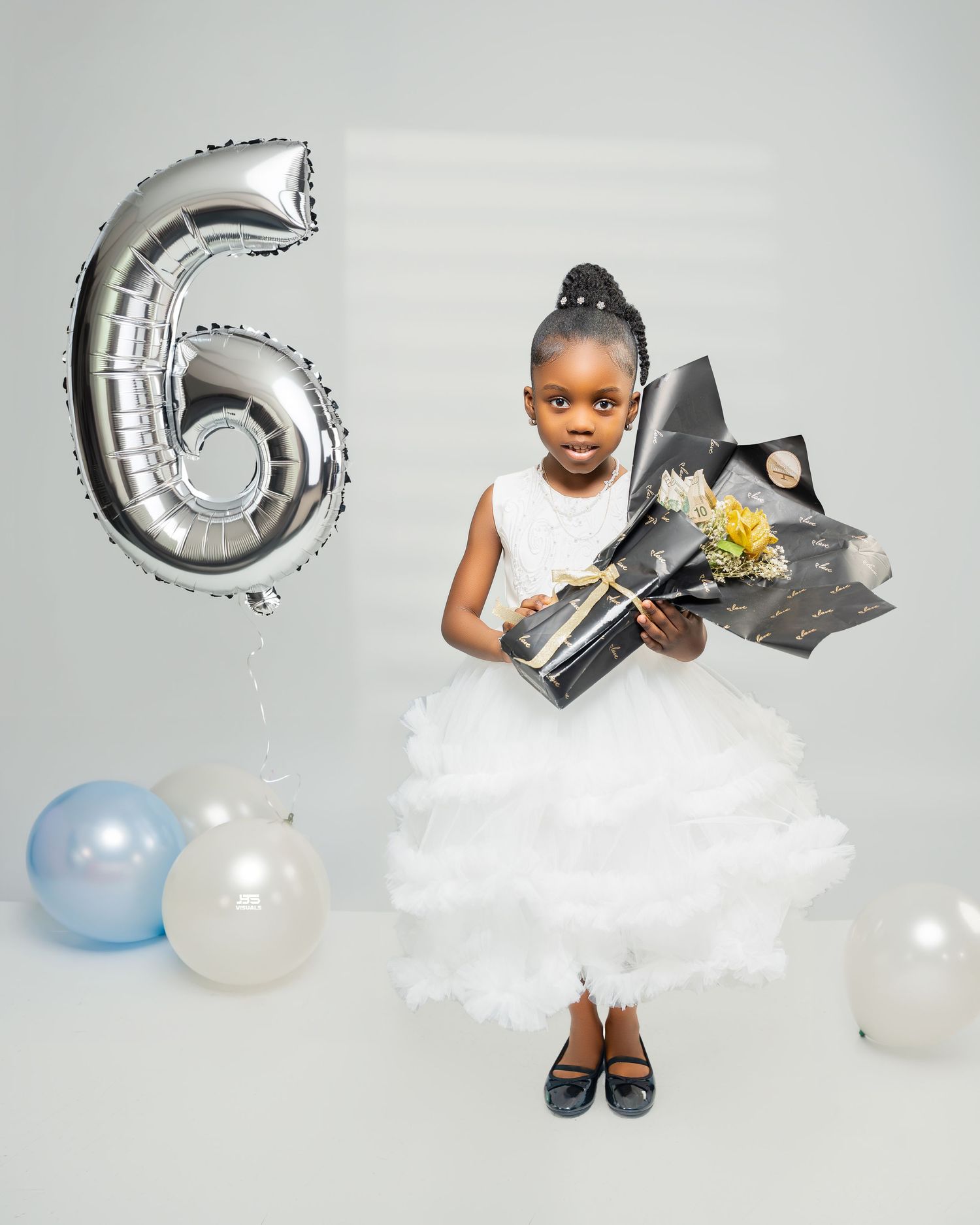 Young girl in white tutu dress holding flowers beside silver number 6 birthday balloon.