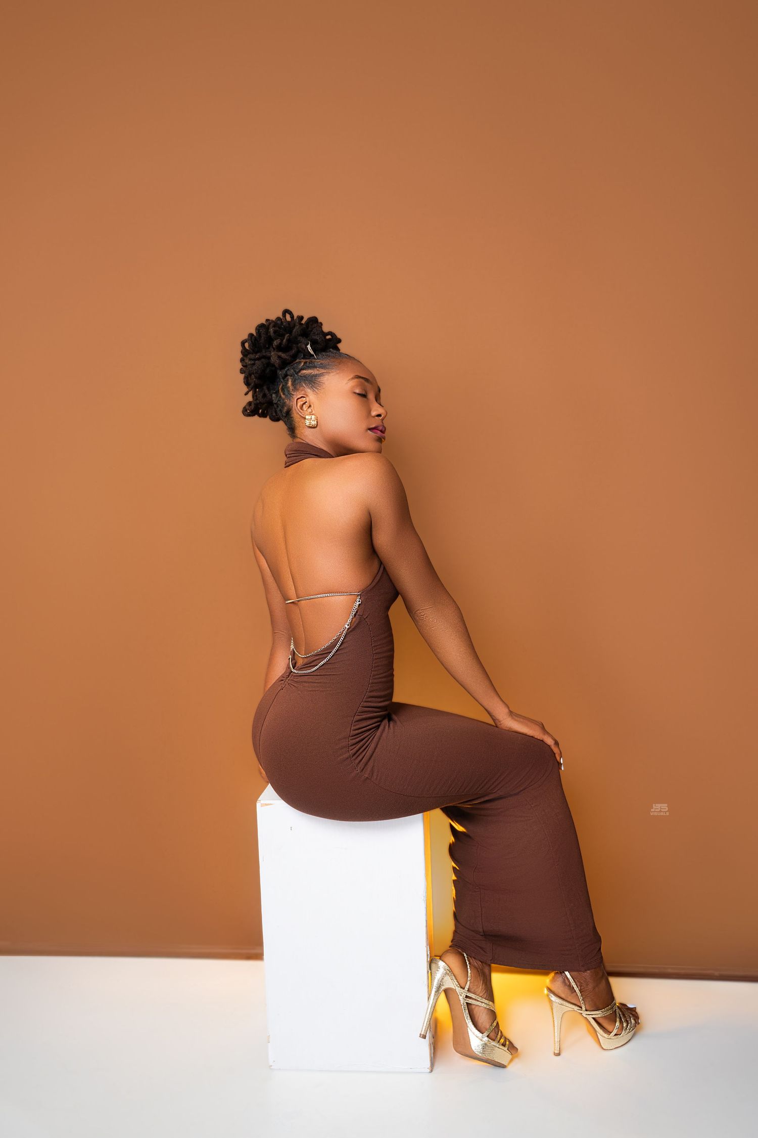 Woman in backless brown halter jumpsuit and gold strappy heels poses seated on white cube, back view.