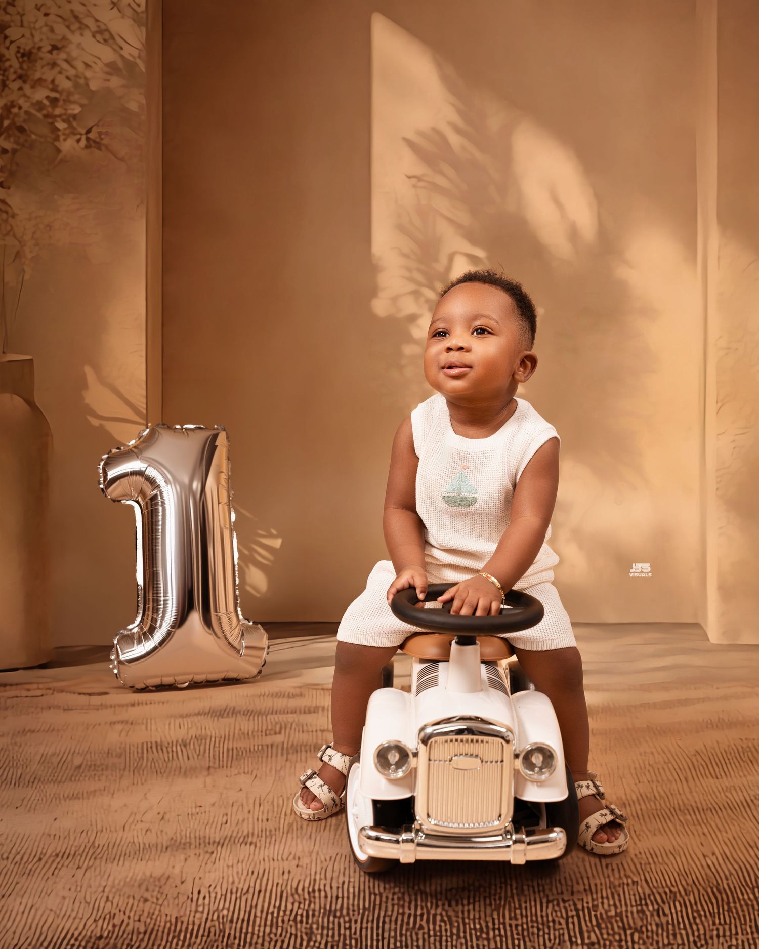 Smiling baby boy sitting on a vintage toy car beside a silver number 1 balloon, celebrating his first birthday.