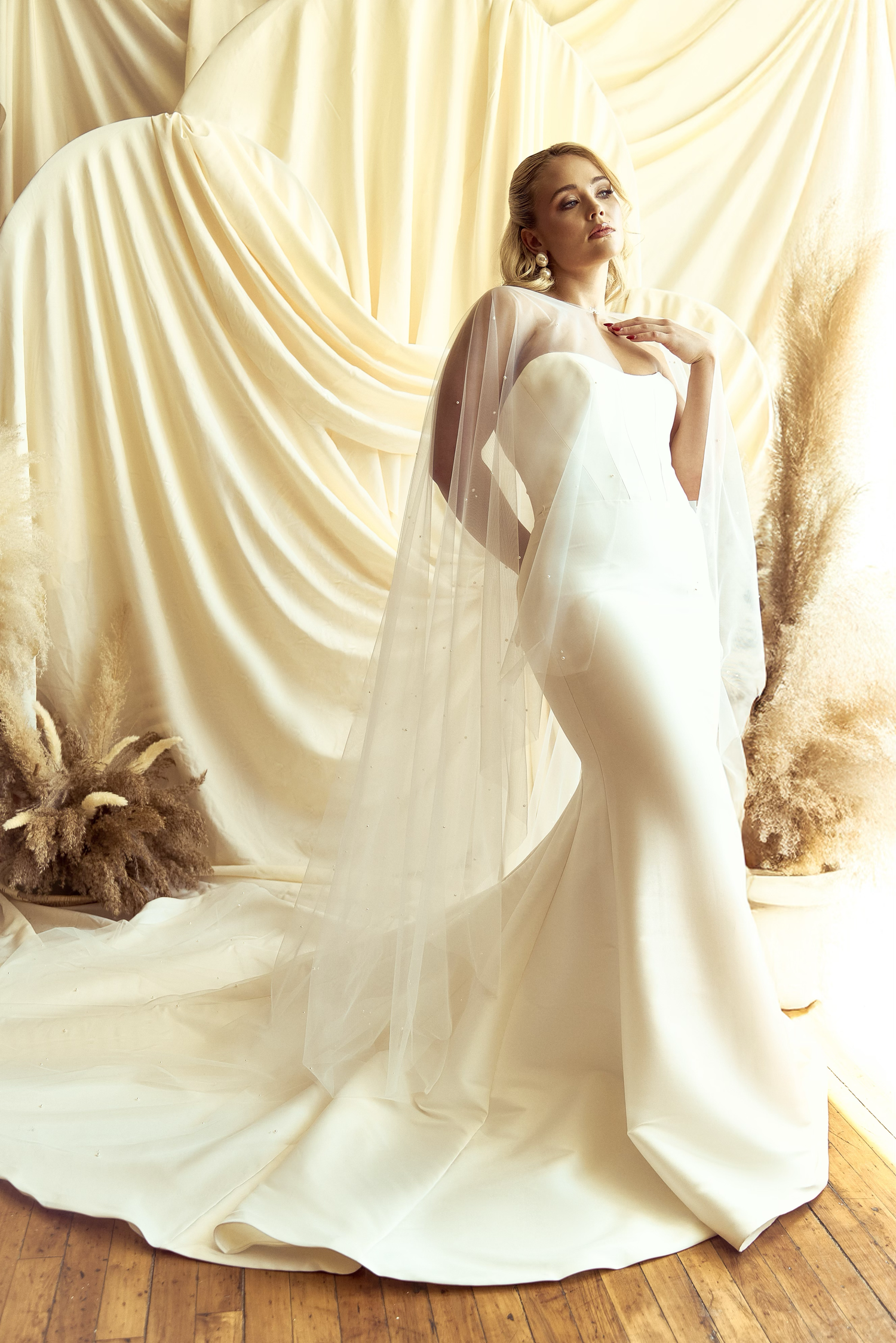 Elegant mermaid-style wedding gown with flowing cape veil against draped cream backdrop with dried pampas grass decor.