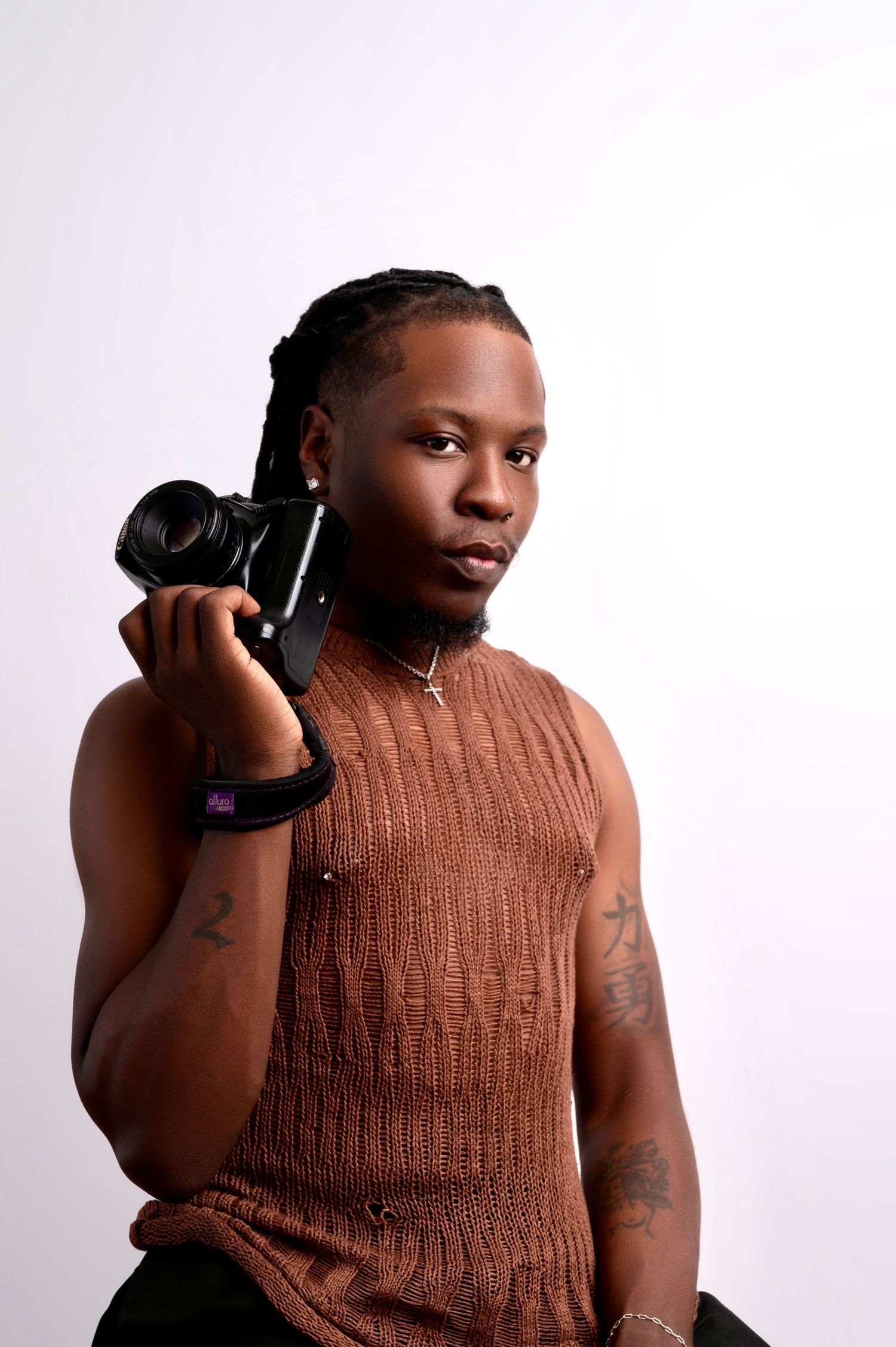 Young Black man with braids holding a camera, wearing a brown sleeveless top and tattoos, posing against white background.
