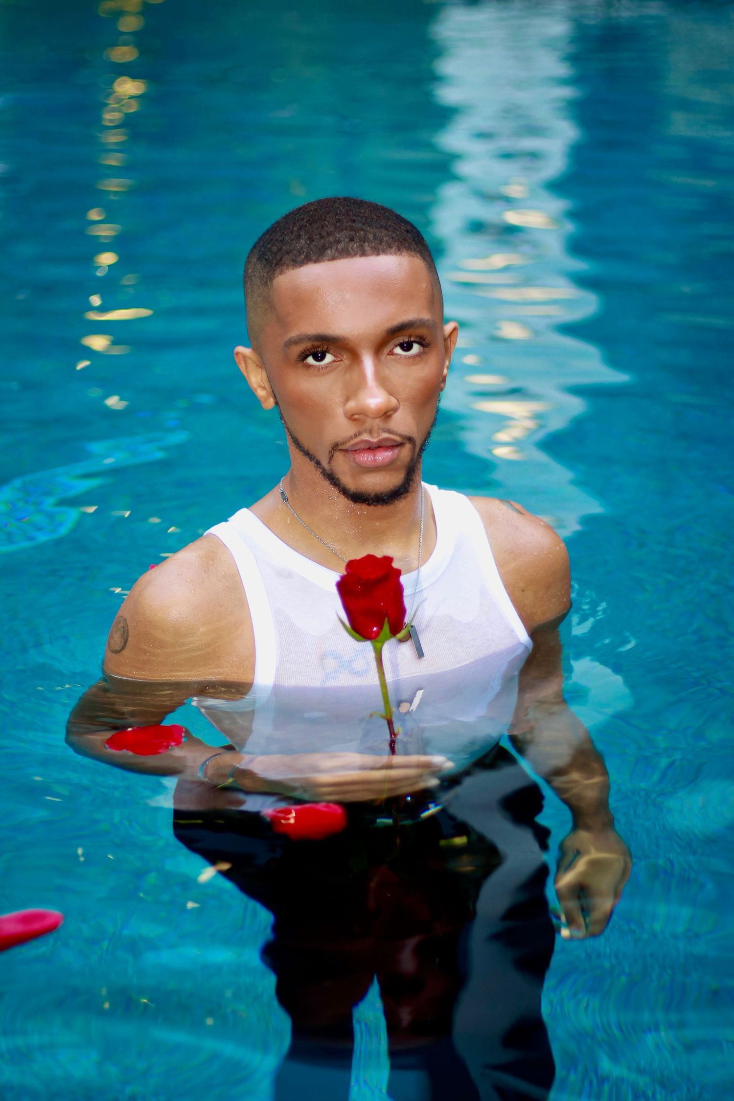 A person stands in turquoise pool water holding a red rose while wearing a white tank top.
