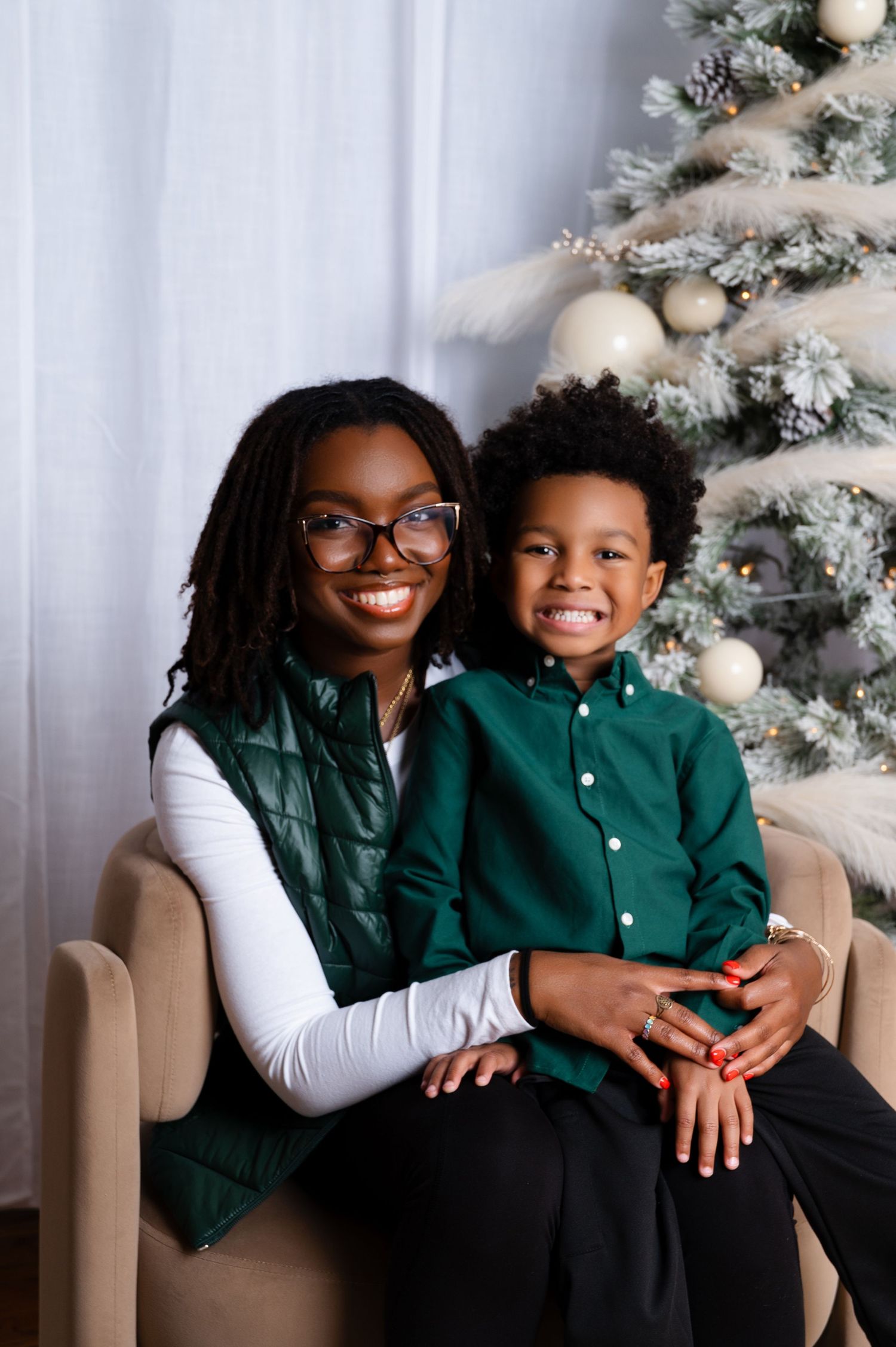 A smiling Black woman in green vest and glasses holds a young boy in green shirt near a decorated Christmas tree.