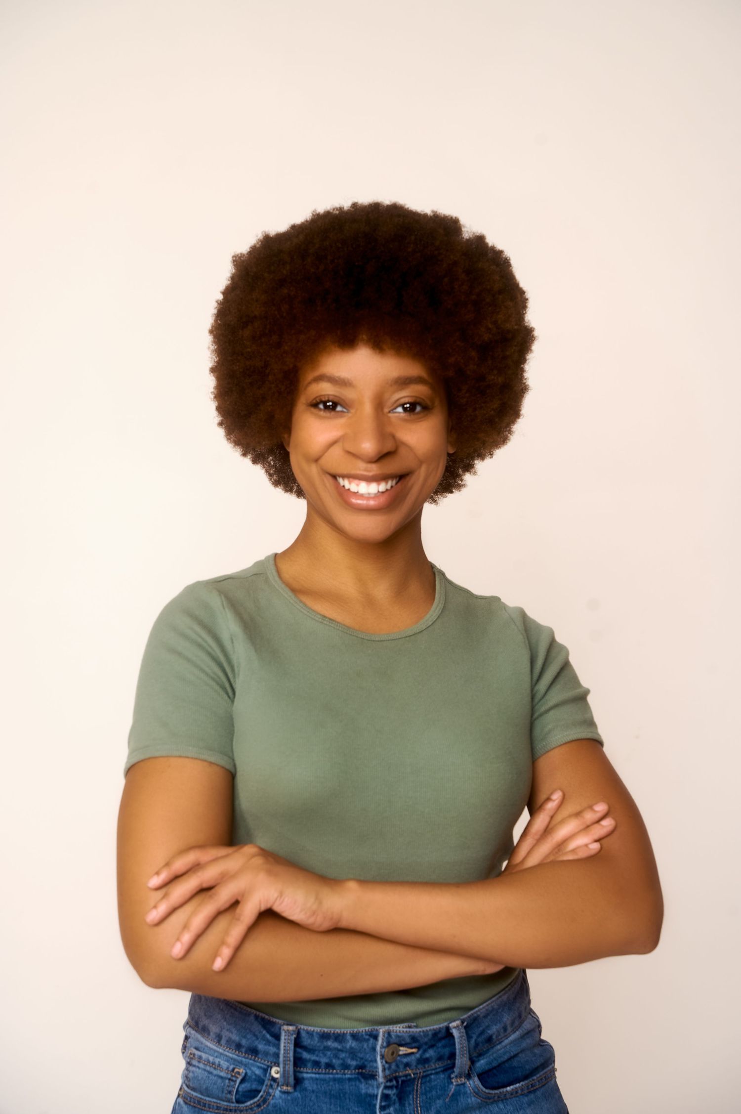 Smiling young woman with afro hair, arms crossed, wearing a sage green t-shirt and jeans against a white background.