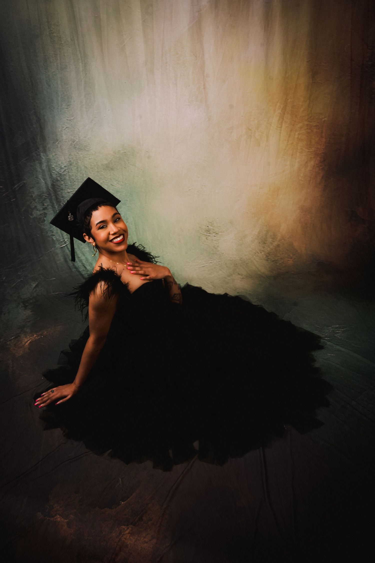 Smiling graduate in black tulle gown and cap poses elegantly on floor against colorful painted backdrop.