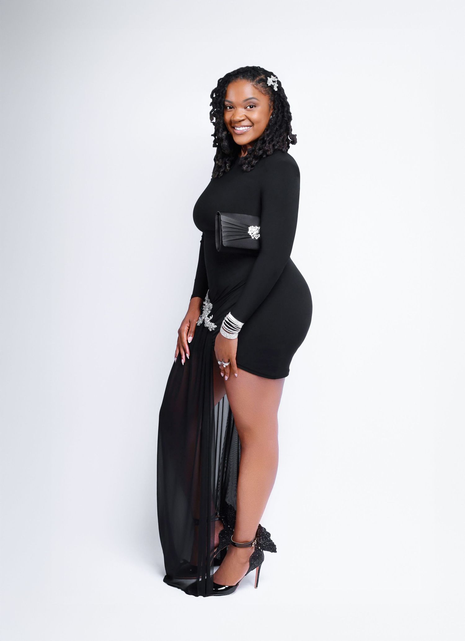 Professional fashion model poses in elegant black dress and heels against white studio backdrop.