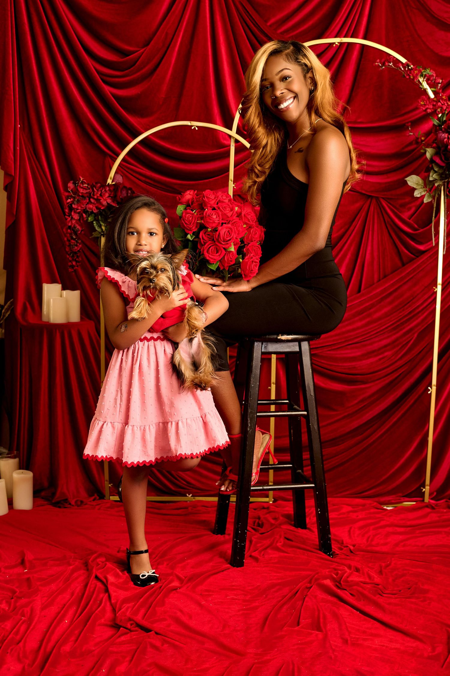 Woman and girl in pink dress pose with Yorkshire Terriers amid red roses and velvet drapes in festive portrait.