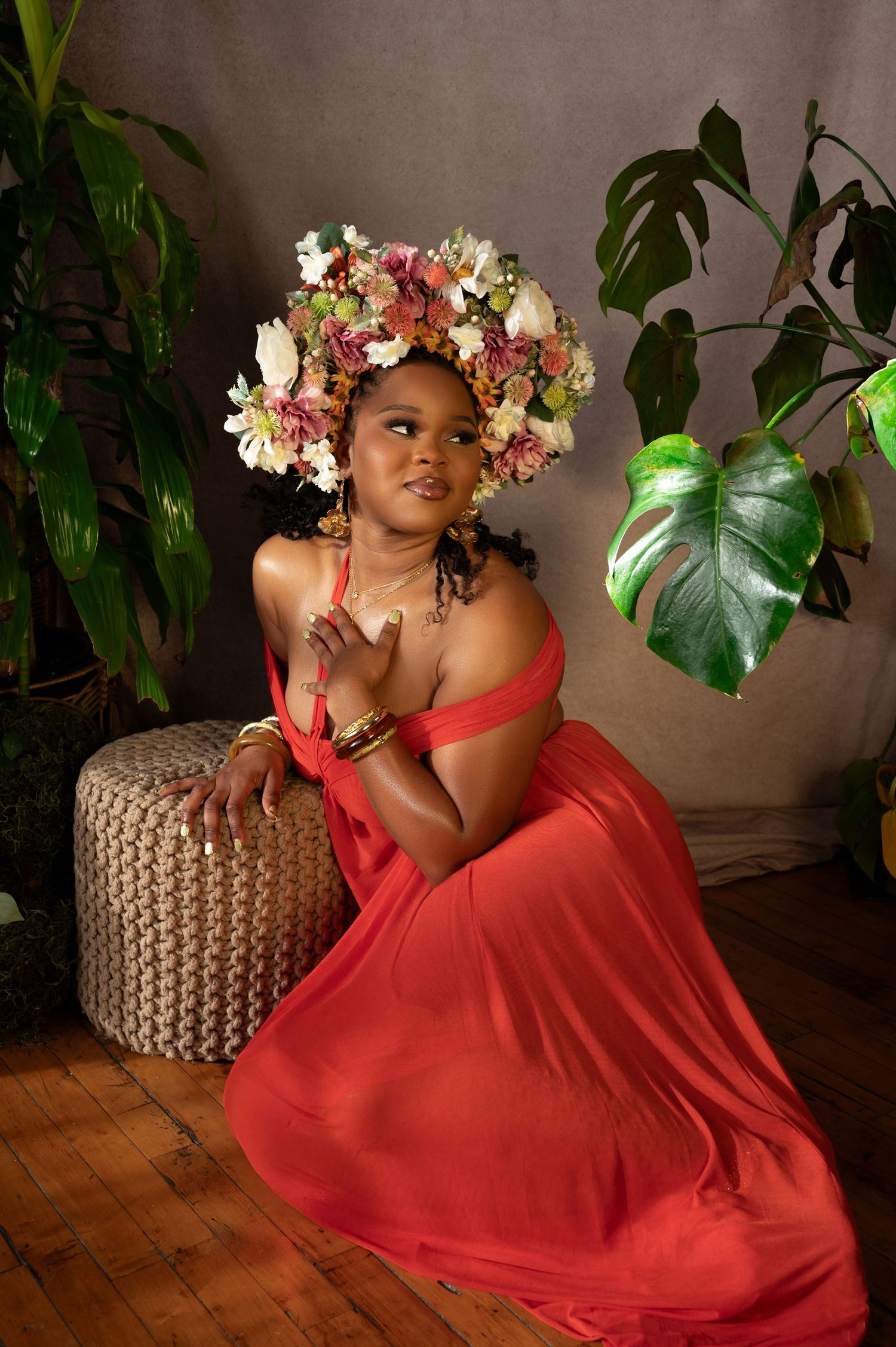 Woman in red dress and floral crown poses gracefully beside tropical plants on wooden floor.