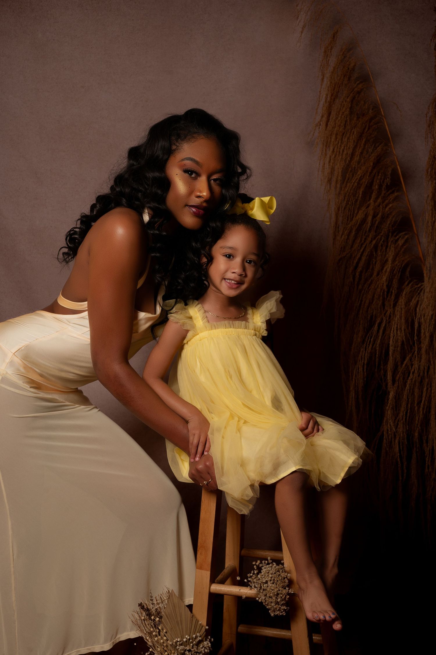 A Black woman in cream dress poses with young girl in yellow tulle dress and bow in studio portrait.