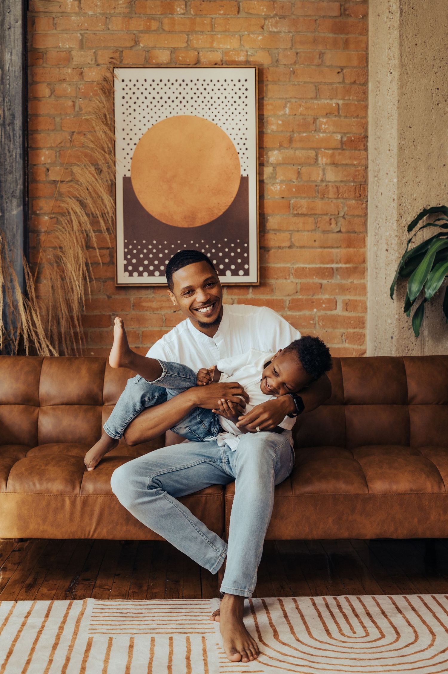 Two people share a joyful moment while sitting on a brown leather couch against an exposed brick wall.