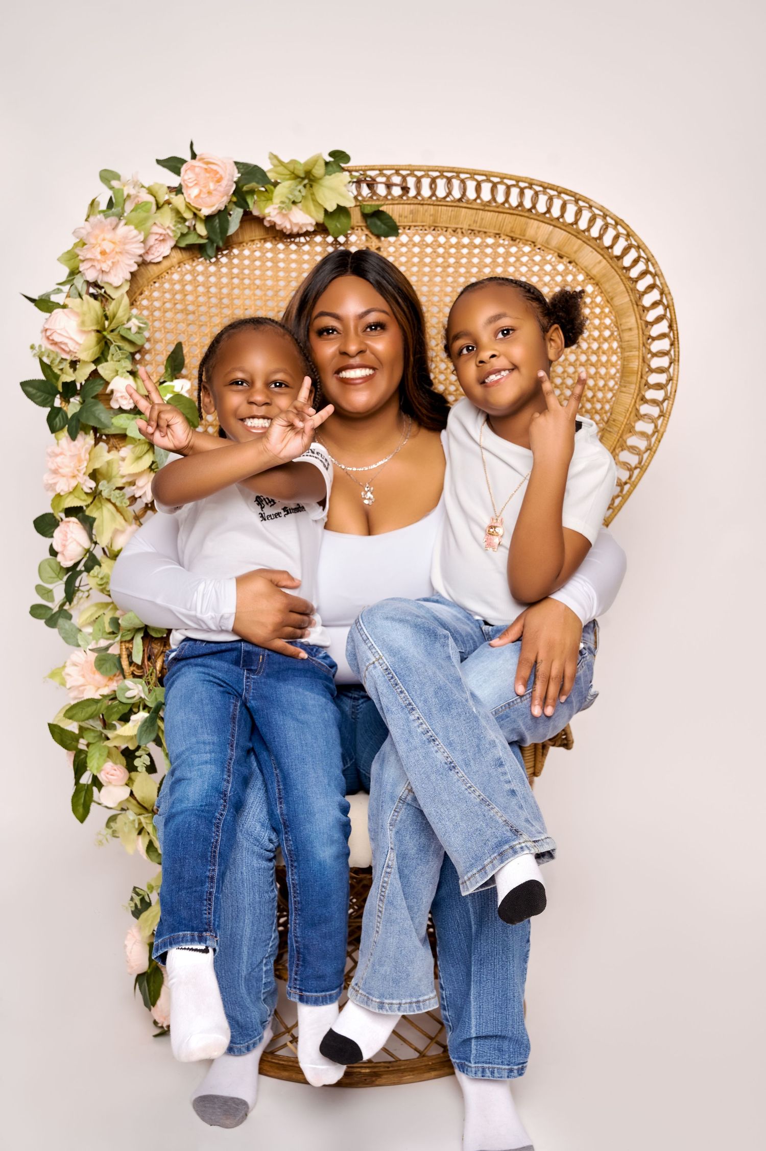 A smiling mother sits in a wicker peacock chair with floral decor, posing with her two children in matching white tops and jeans.