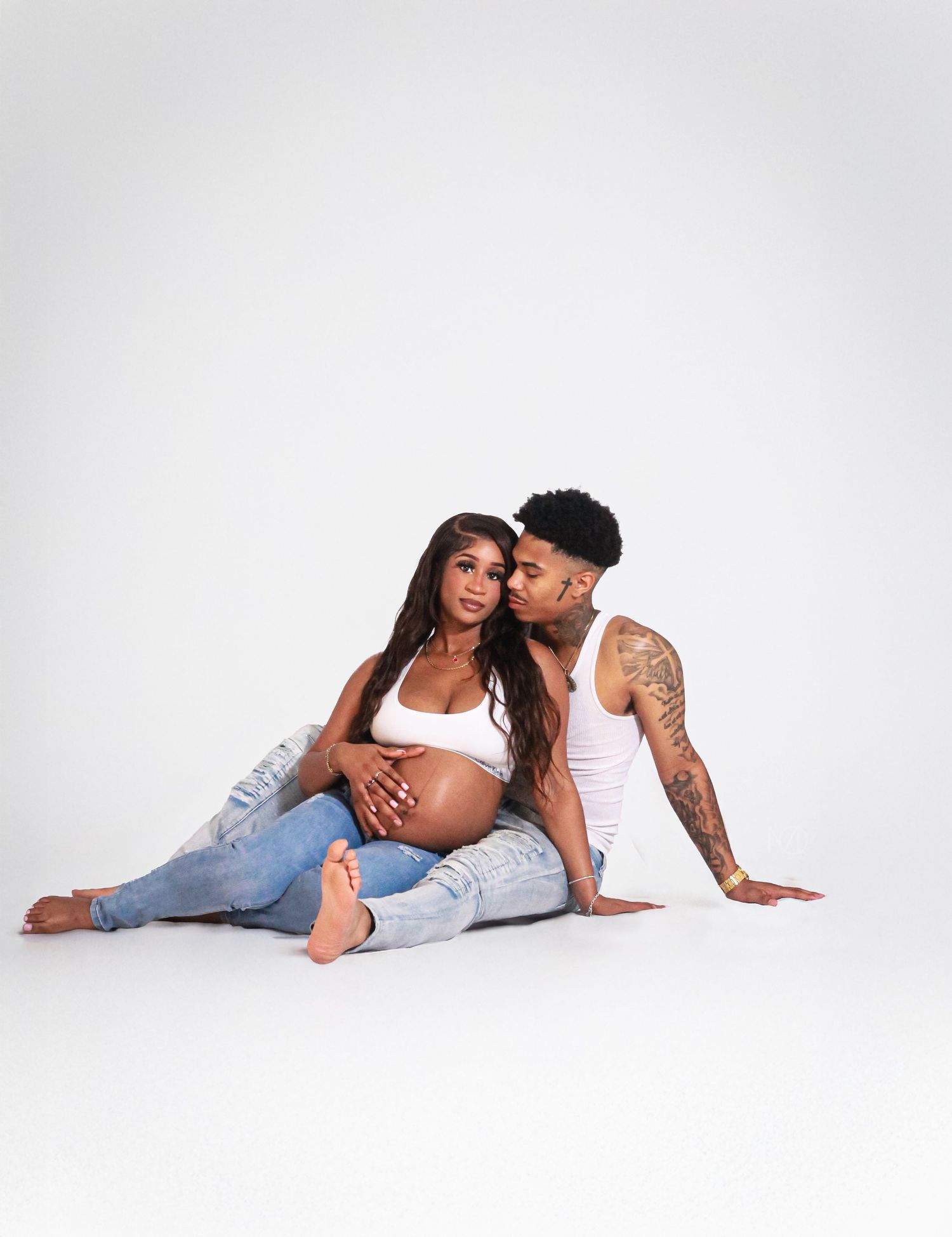 A couple in casual white and denim outfits pose intimately together on the floor against a white background.