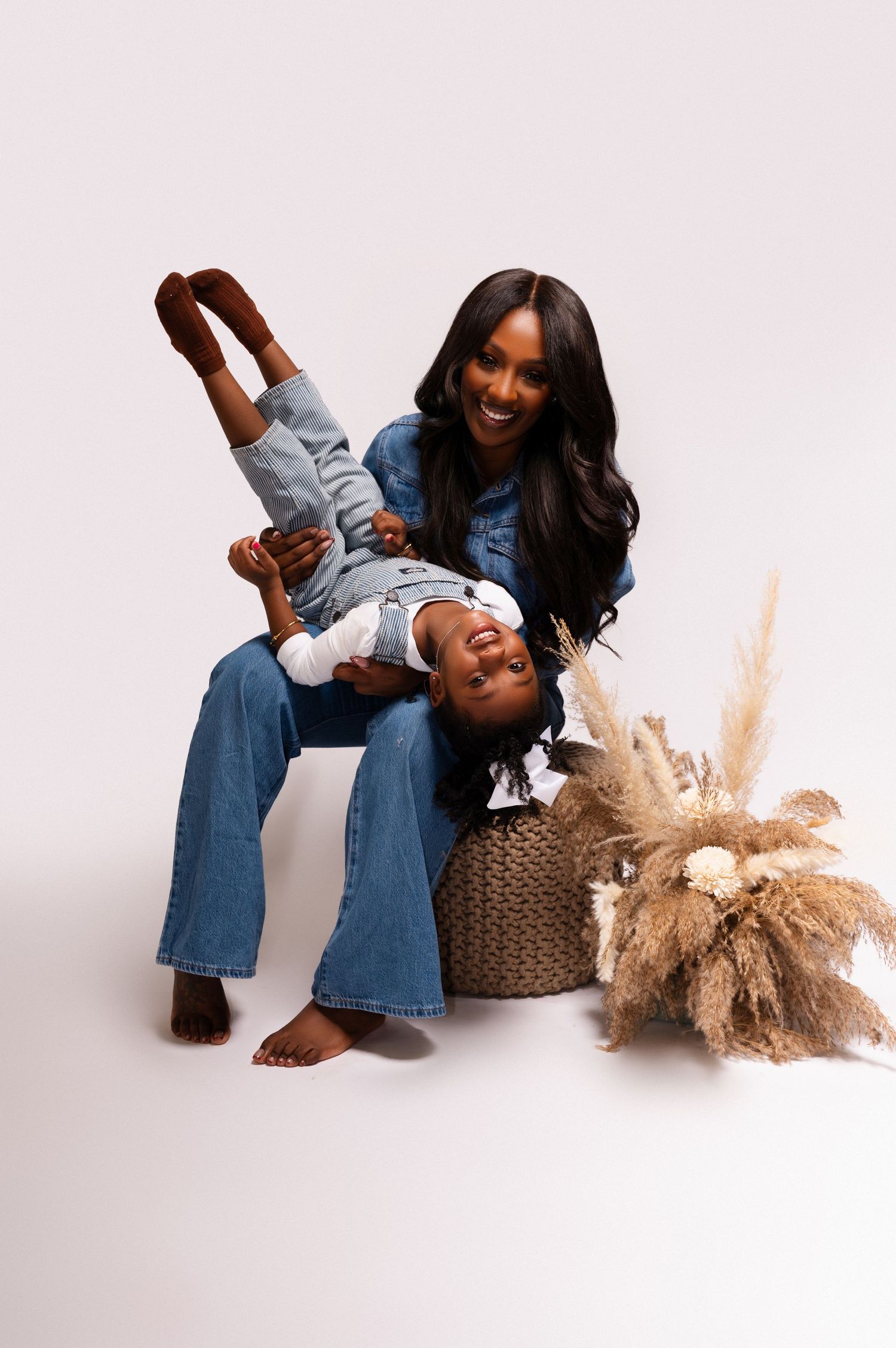 A joyful mother in denim holds her laughing child upside down beside a woven basket with pampas grass.
