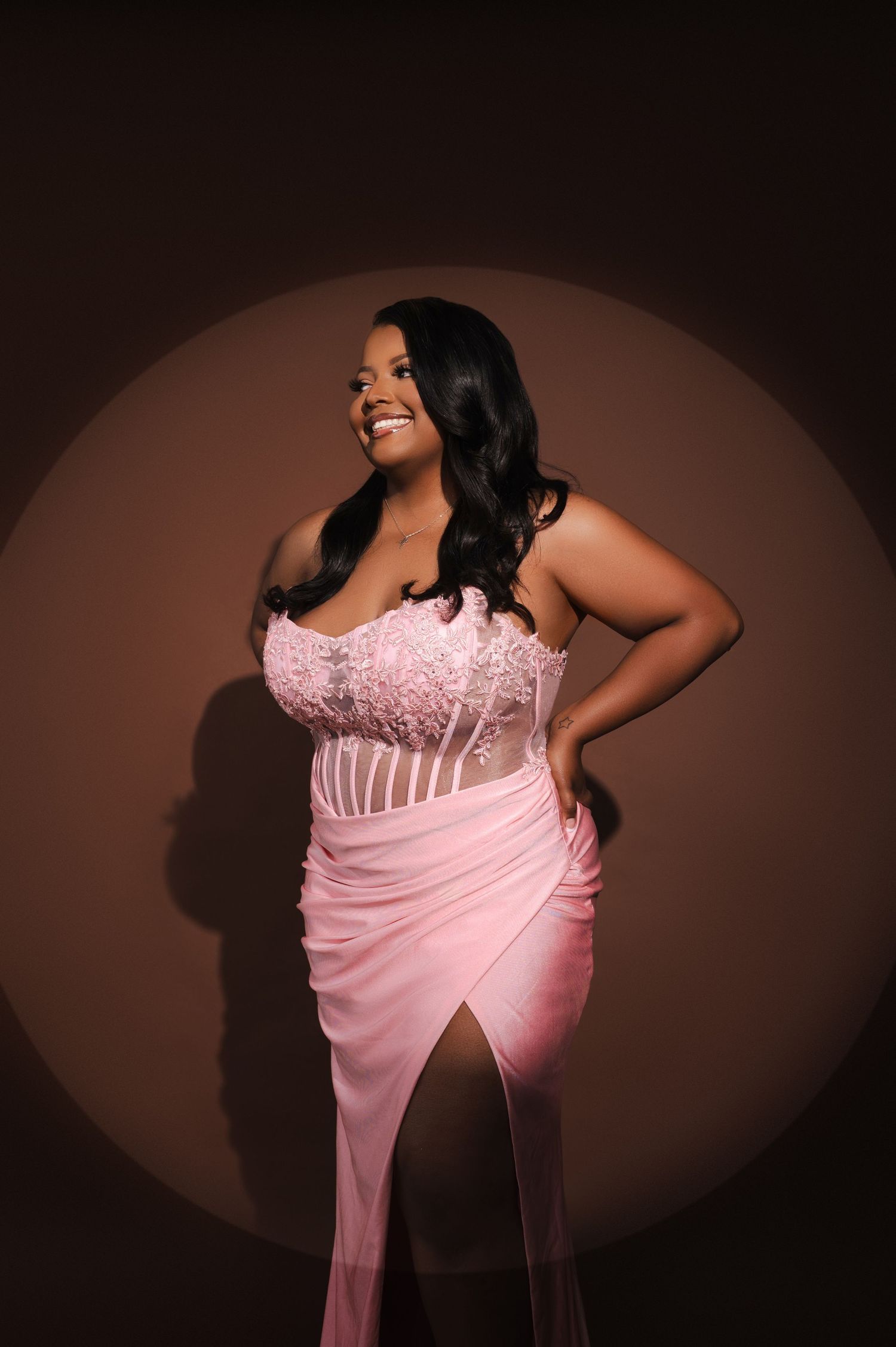 A smiling woman in a pink strapless gown with floral details and a slit poses confidently against a brown backdrop.