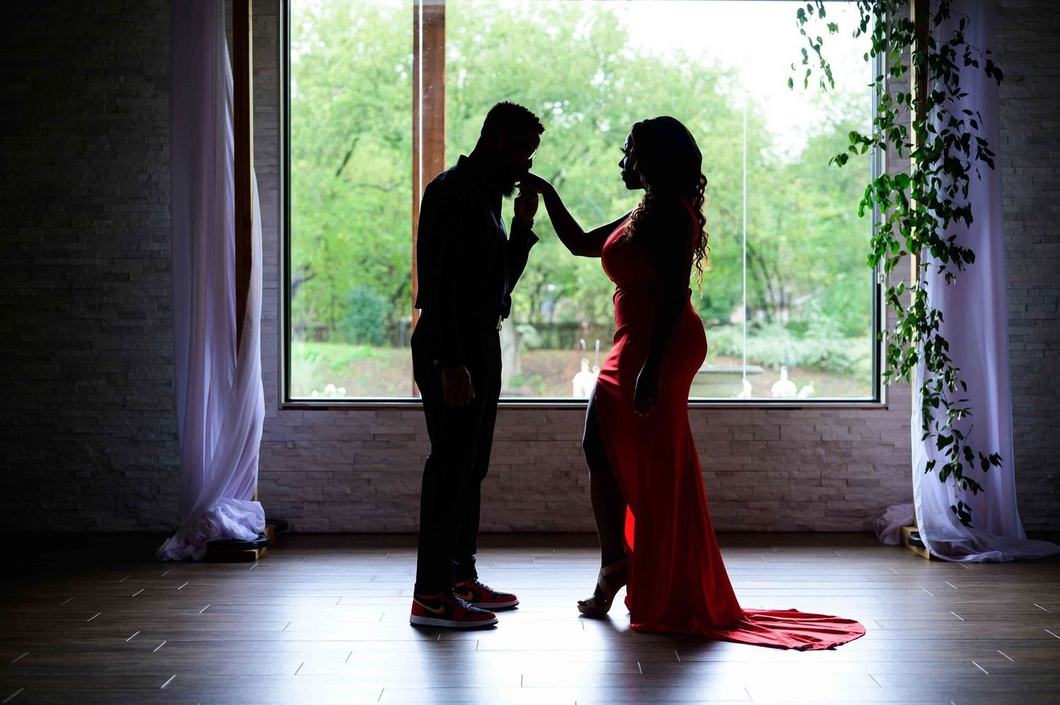 Silhouette of a couple sharing a romantic moment by a large window, woman in red dress, lush greenery outside.