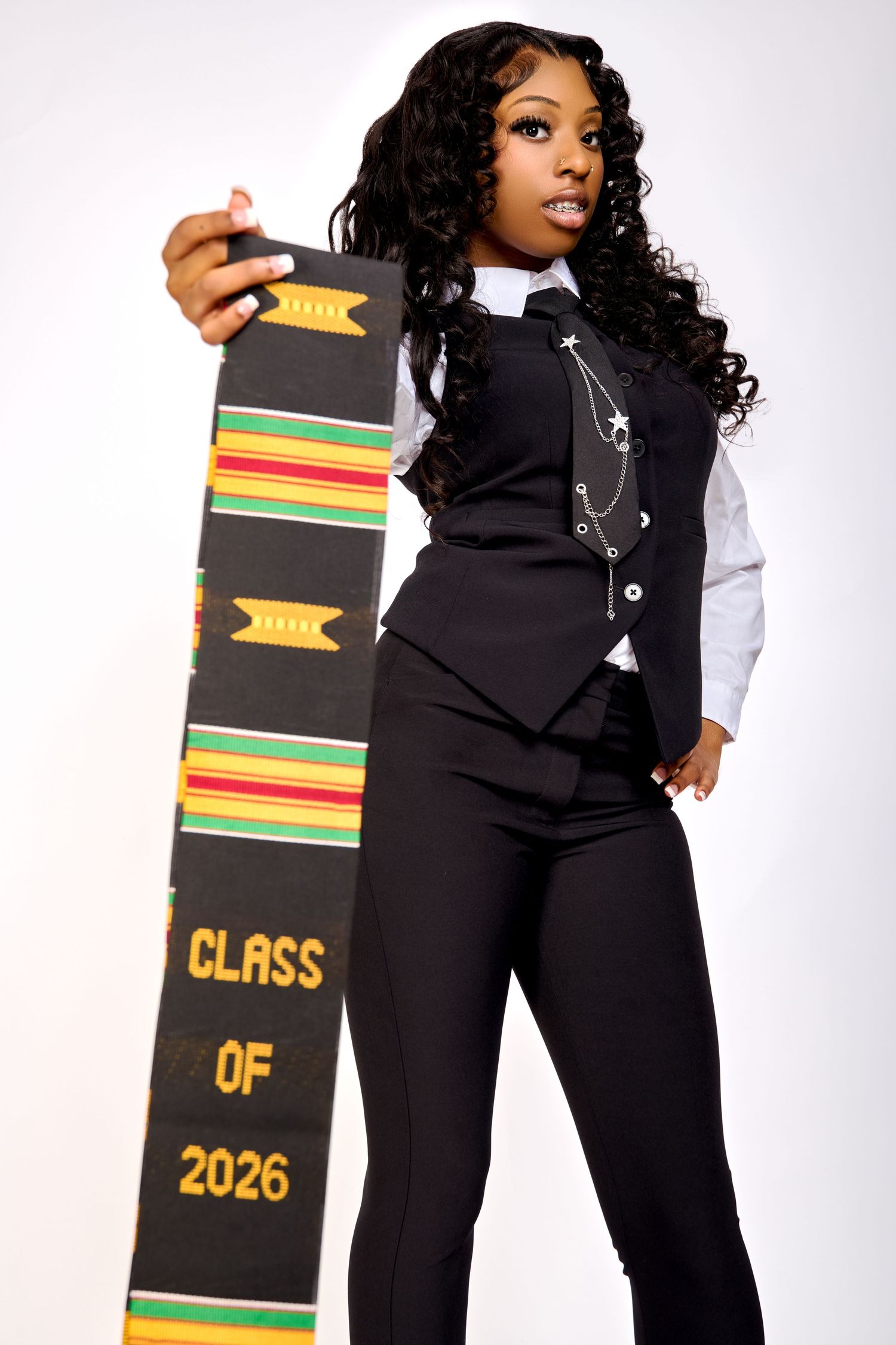 Young woman in black vest holding a Kente cloth graduation stole reading Class of 2026 against white background.
