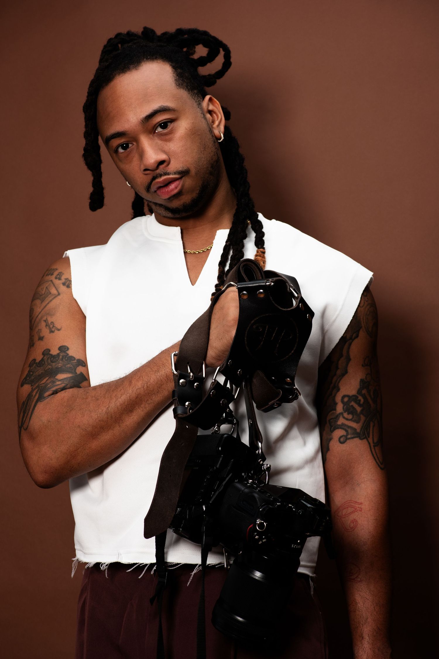 A person in a white sleeveless shirt poses with a camera against a brown backdrop in a professional photo series.