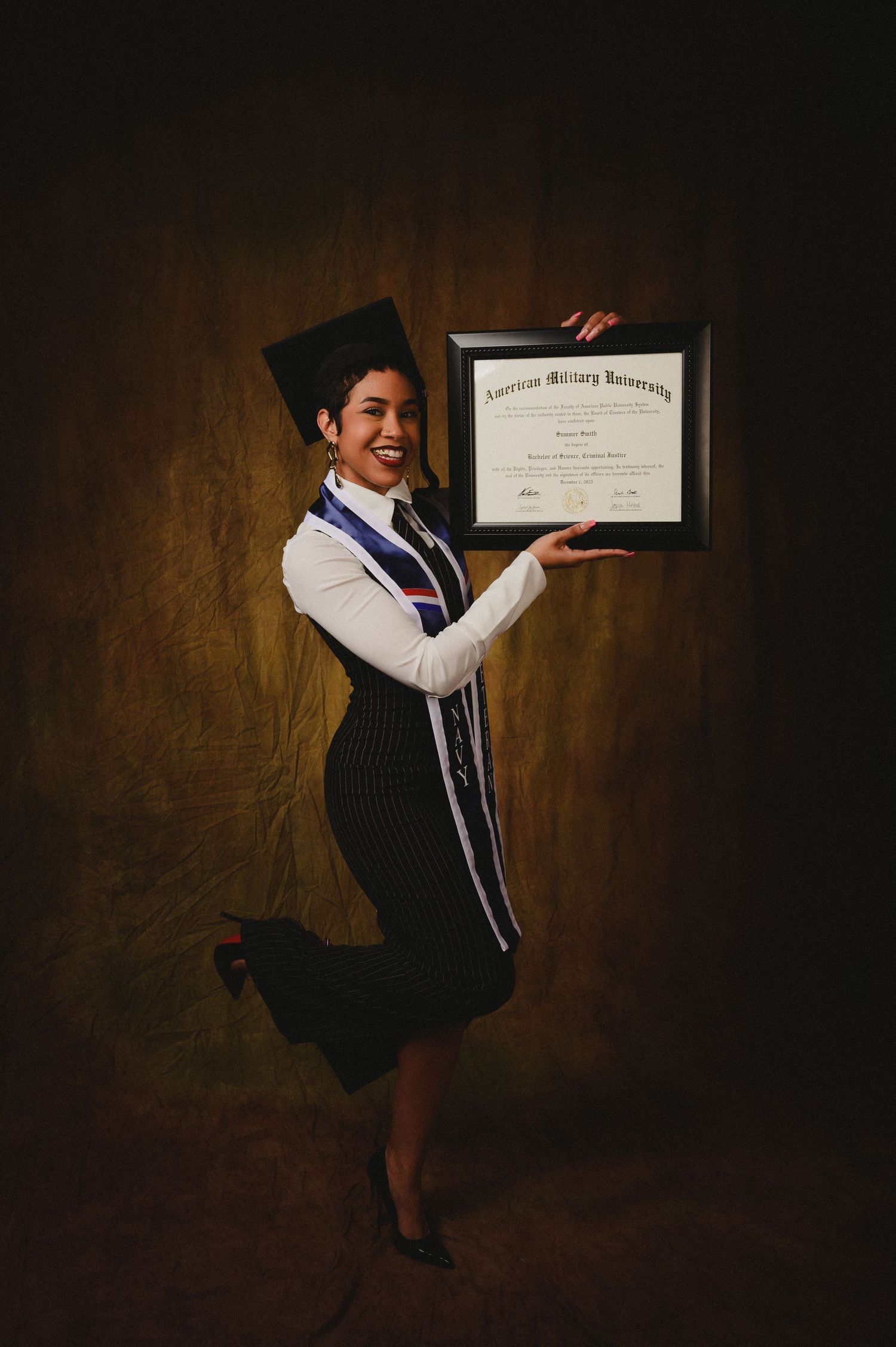 Smiling graduate in cap and gown holds American Military University diploma, posing joyfully against dark background.