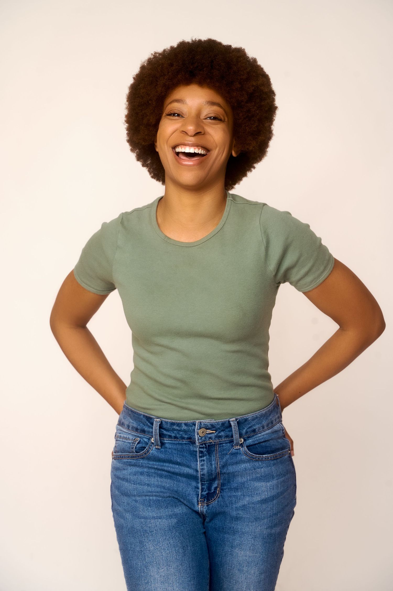 Smiling young Black woman with afro hair wearing sage green t-shirt and blue jeans, hands on hips, white background.