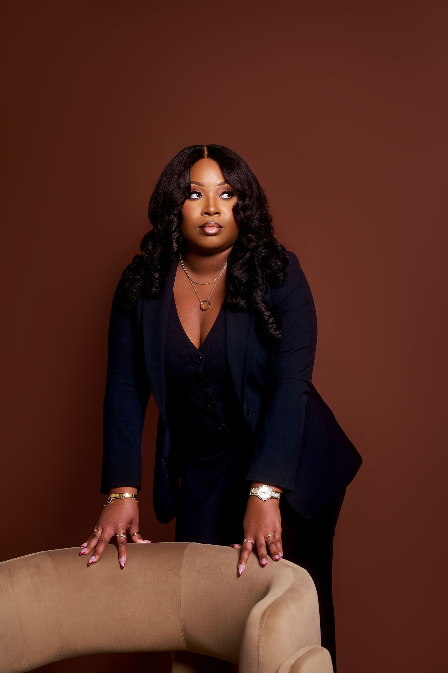 A confident Black woman in a navy blazer poses leaning on a beige chair against a brown studio background.