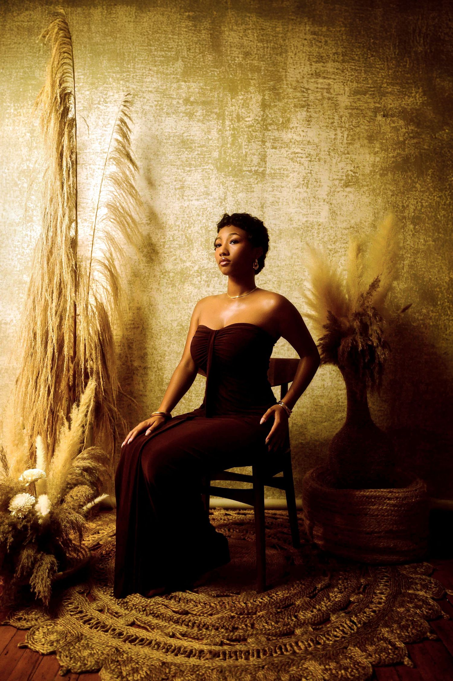 Artistic portrait in burgundy gown against golden wall with dried pampas grass decor.