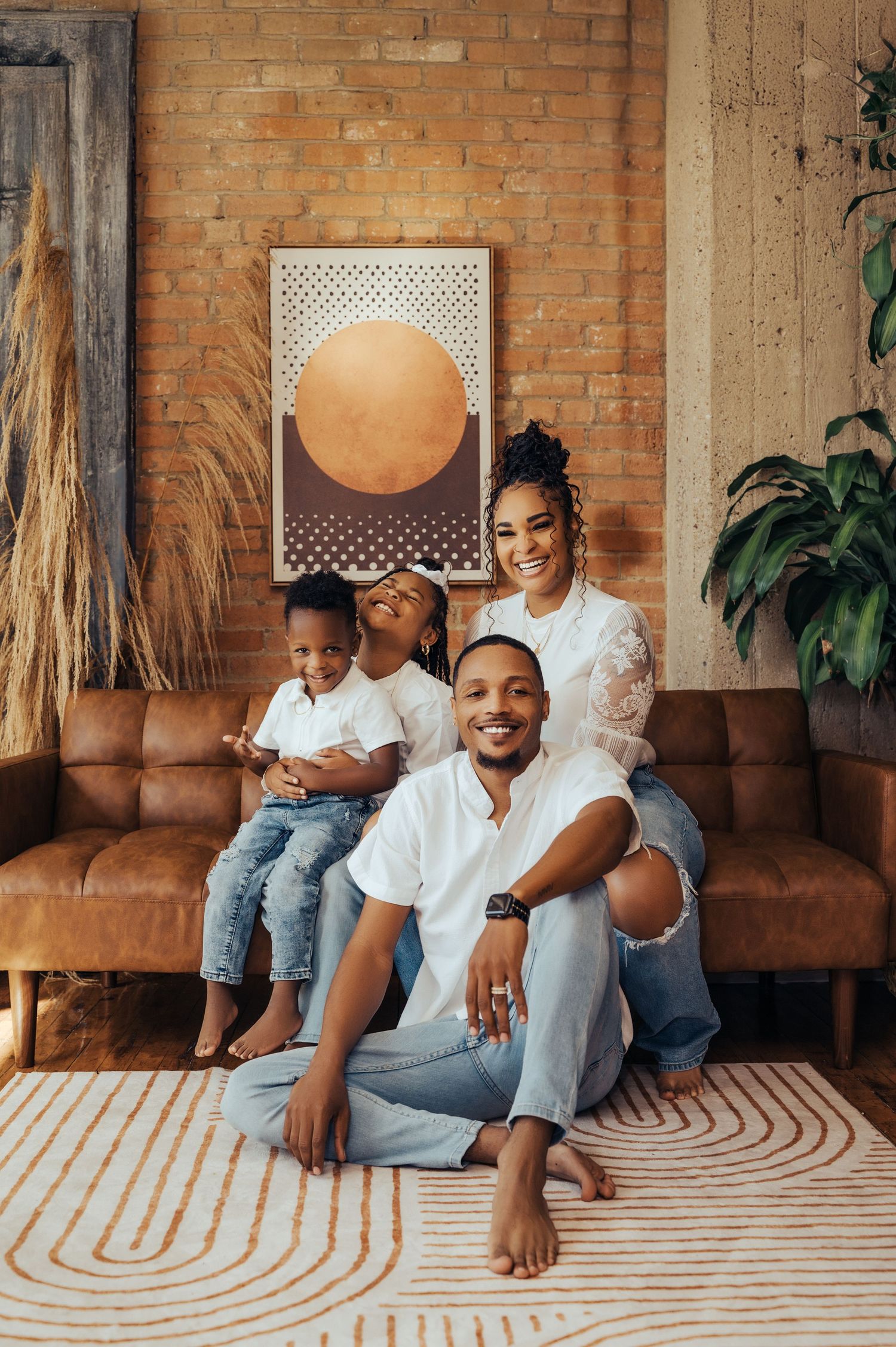 Family relaxing on patterned rug against exposed brick wall in casual white and denim outfits.