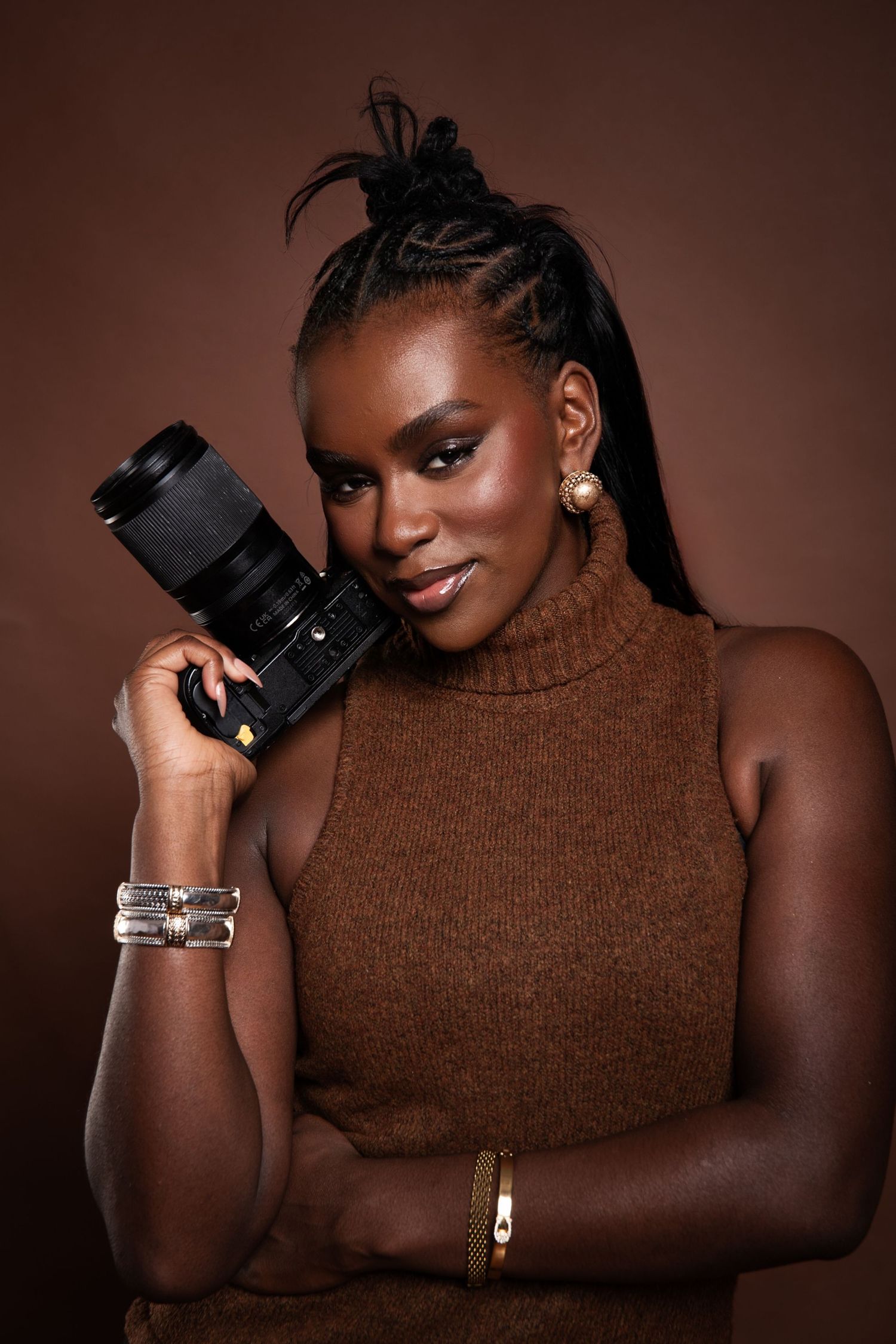 Professional photographer in brown turtleneck sweater poses with camera against dark brown background.