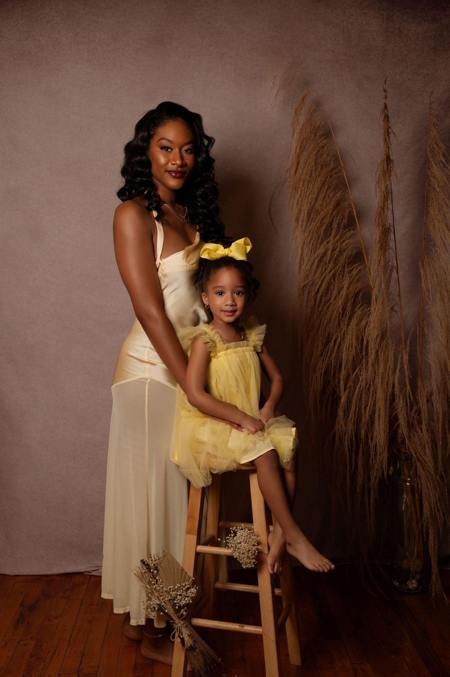 A woman in a cream gown poses with a young girl in a yellow tutu dress and bow on a wooden stool in a studio.