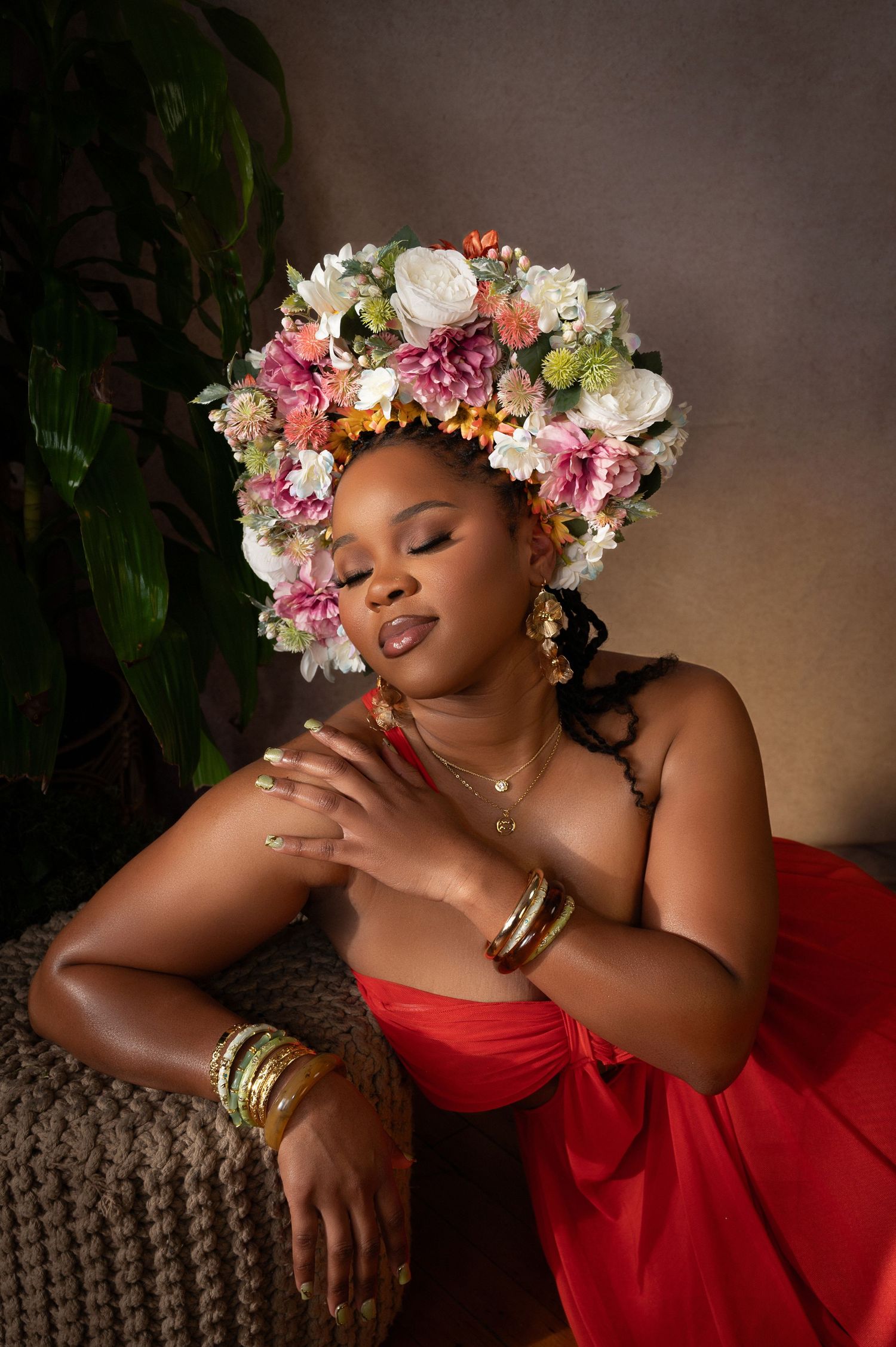 Woman in red dress wearing colorful floral crown and gold jewelry, posing gracefully with eyes closed near tropical plants.